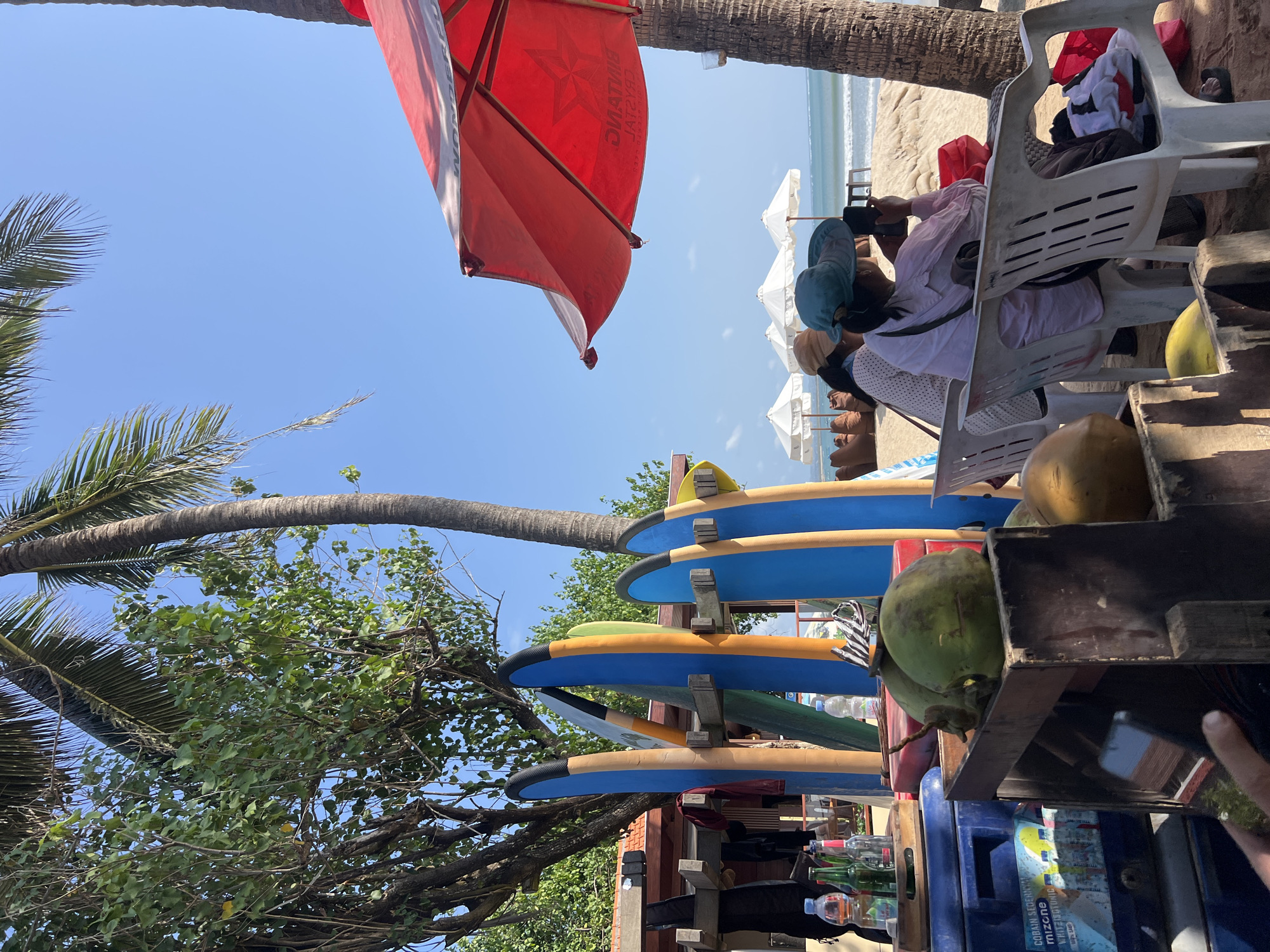 The image depicts a beach scene in Kuta, Indonesia, on a clear, sunny day. In the foreground, there are several surfboards leaning against a wooden rack. Next to them, on a blue cooler, are bottles of drinks and coconuts. Further back, three people are seated on chairs, facing the ocean. One person is wearing a blue hat and holding a phone. The beach is sandy, and in the distance, there are white umbrellas and lounge chairs. Palm trees are visible throughout the scene, and a red umbrella is prominent on the right side of the image. The overall atmosphere suggests a relaxed beach setting.