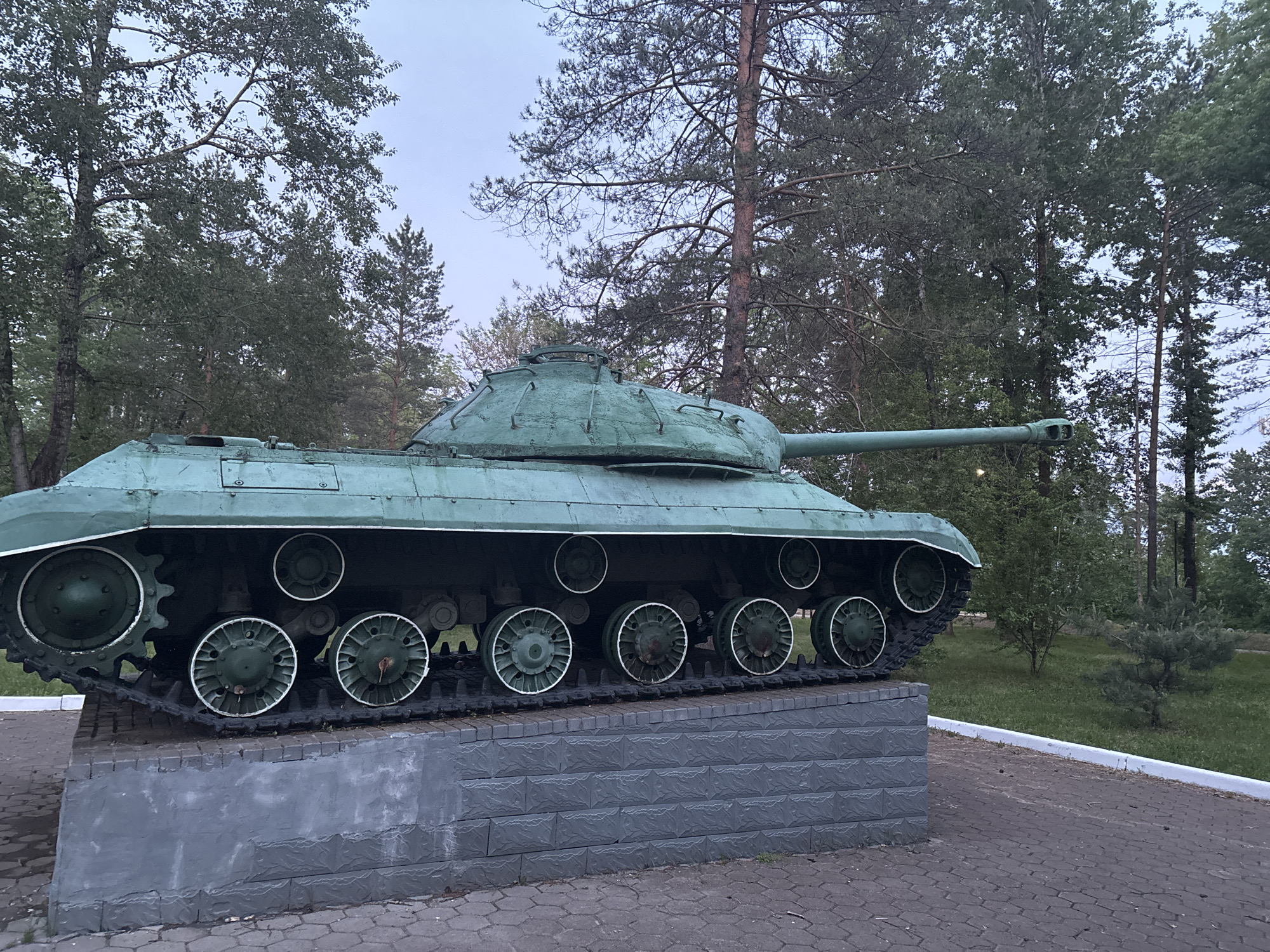 The image displays a large, dark green tank, likely a historical monument, positioned outdoors on a stone pedestal. The tank is painted in a faded, mossy green, with lighter green or white accents on the edges of its track skirts and road wheels. Its long gun barrel extends towards the right, while a rounded turret with visible hatches sits atop the hull. The tank's track system and numerous spoked road wheels are clearly visible, contrasting with the dark tracks.
The tank rests on a rectangular base constructed from grey, block-patterned stones. The ground around the base is paved with grey hexagonal or octagonal interlocking stones, and a low, light-colored curb delineates the paved area from a grassy patch.
The setting is a park-like or forested environment. Tall, leafy trees, including both broadleaf and coniferous varieties, form a dense green background, reaching up towards an overcast, light grey sky. The lighting is soft and diffuse, suggesting an early morning or late afternoon without direct sunlight. No people are present in the scene, and there is no discernible text on the tank or its surroundings. The overall impression is one of a static display, possibly a war memorial or museum exhibit.