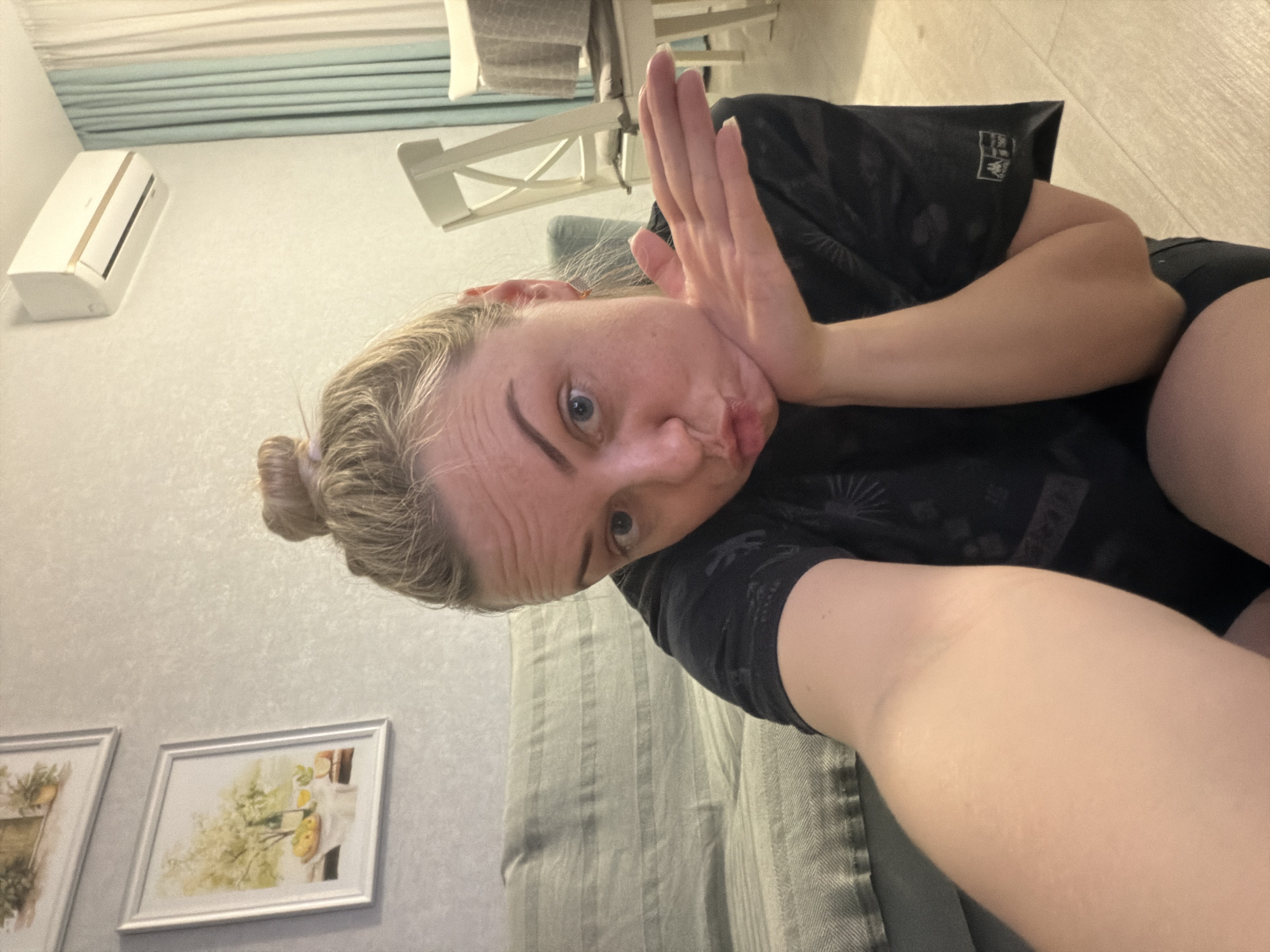 A woman is taking a selfie indoors. She is lying on a gray striped couch, wearing a black t-shirt. Her hair is tied in a bun. She is holding her hand up near her face, making a pouting expression. In the background are framed pictures on the wall, an air conditioner, curtains, and a white chair. The location is Votkinsk, Russia.