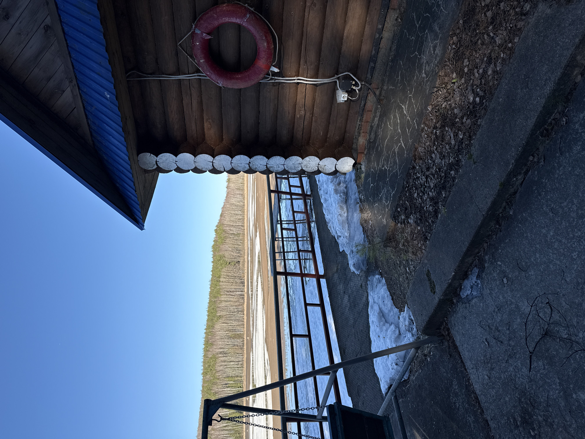 The image depicts a lakeside scene in Chaykovsky, Russia. A wooden structure with a blue roof overhangs a cracked concrete platform. A red life preserver is mounted to the wooden ceiling of the structure. A metal ladder leads from the platform into a partially frozen lake. A forest lines the opposite shore under a clear blue sky. Patches of snow remain on the platform near the ladder.