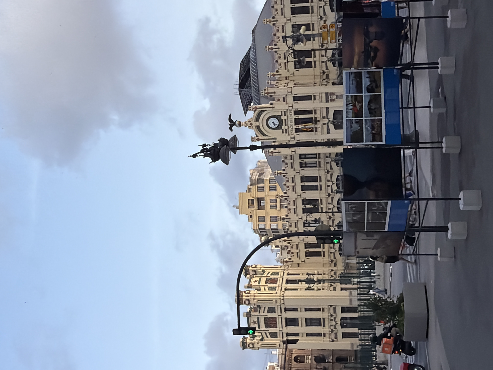 The image depicts an outdoor exhibition in front of a grand, cream-colored building with ornate architecture. The building features multiple stories, arched windows, decorative stonework, and what appears to be a clock face and a flag. A street lamp with a nautical-themed lantern stands prominently in the foreground, partially obscuring the building. To the left of the building, a traffic light displays a green arrow.
In the foreground, a series of framed photographs are displayed on black metal stands, each resting on concrete bases. Some of these photographs appear to be landscapes or scenic views. A large concrete planter with greenery is situated on the left side of the street. A person on a red scooter with an orange delivery box on the back is visible. In the background, several pedestrians are walking on the sidewalk. The sky above is a light blue with scattered clouds. The overall scene suggests a public space, possibly a plaza or street, in front of a historic or significant building.