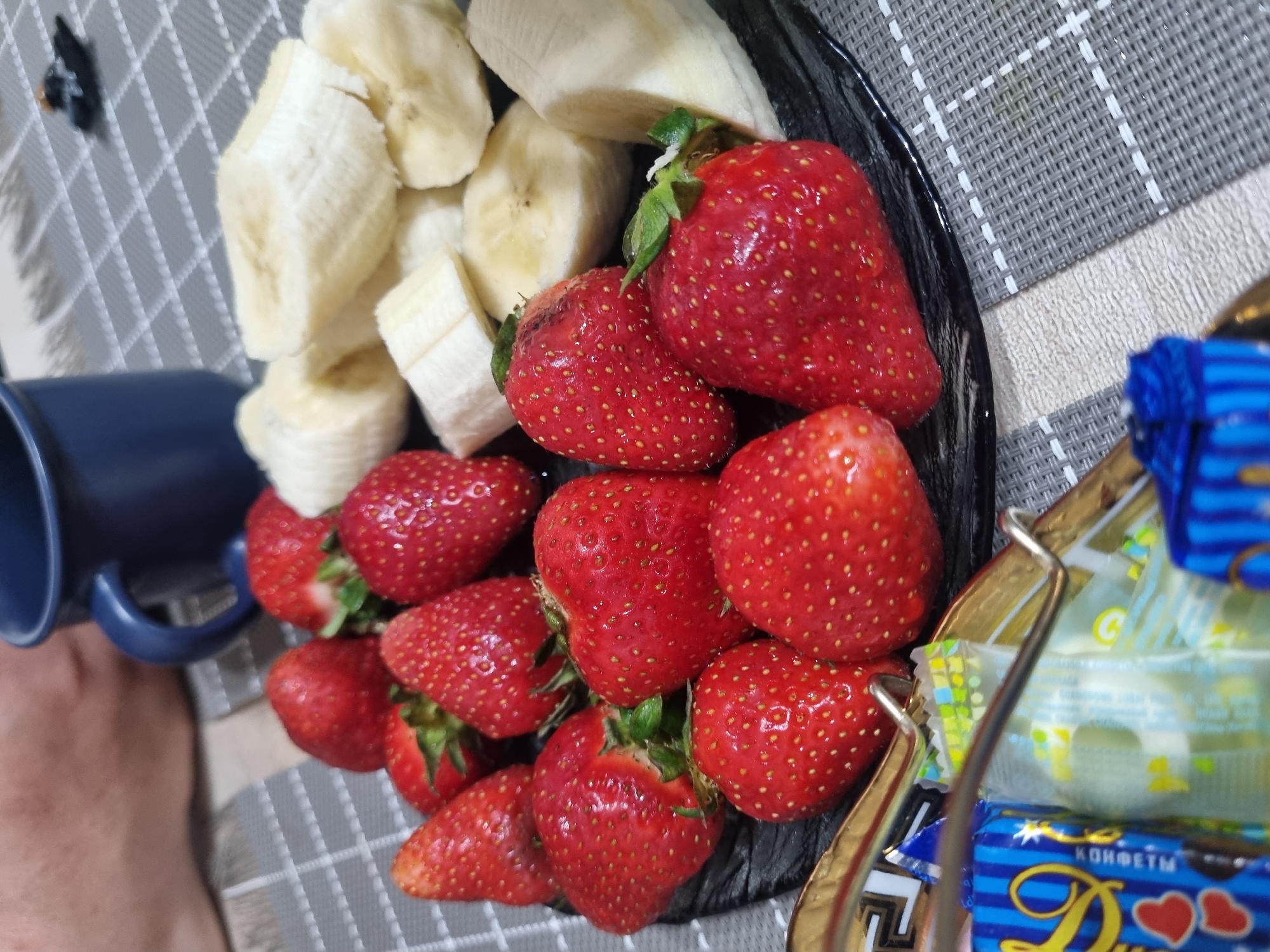 On a table with a gray and white woven pattern cloth is a black plate containing sliced bananas and strawberries. A blue mug is positioned to the left of the plate. To the right of the plate is a multi-tiered metal confectionery stand with wrapped candies. The image does not contain enough information to determine if it was taken in Sochi, Russia. The image primarily depicts food items.