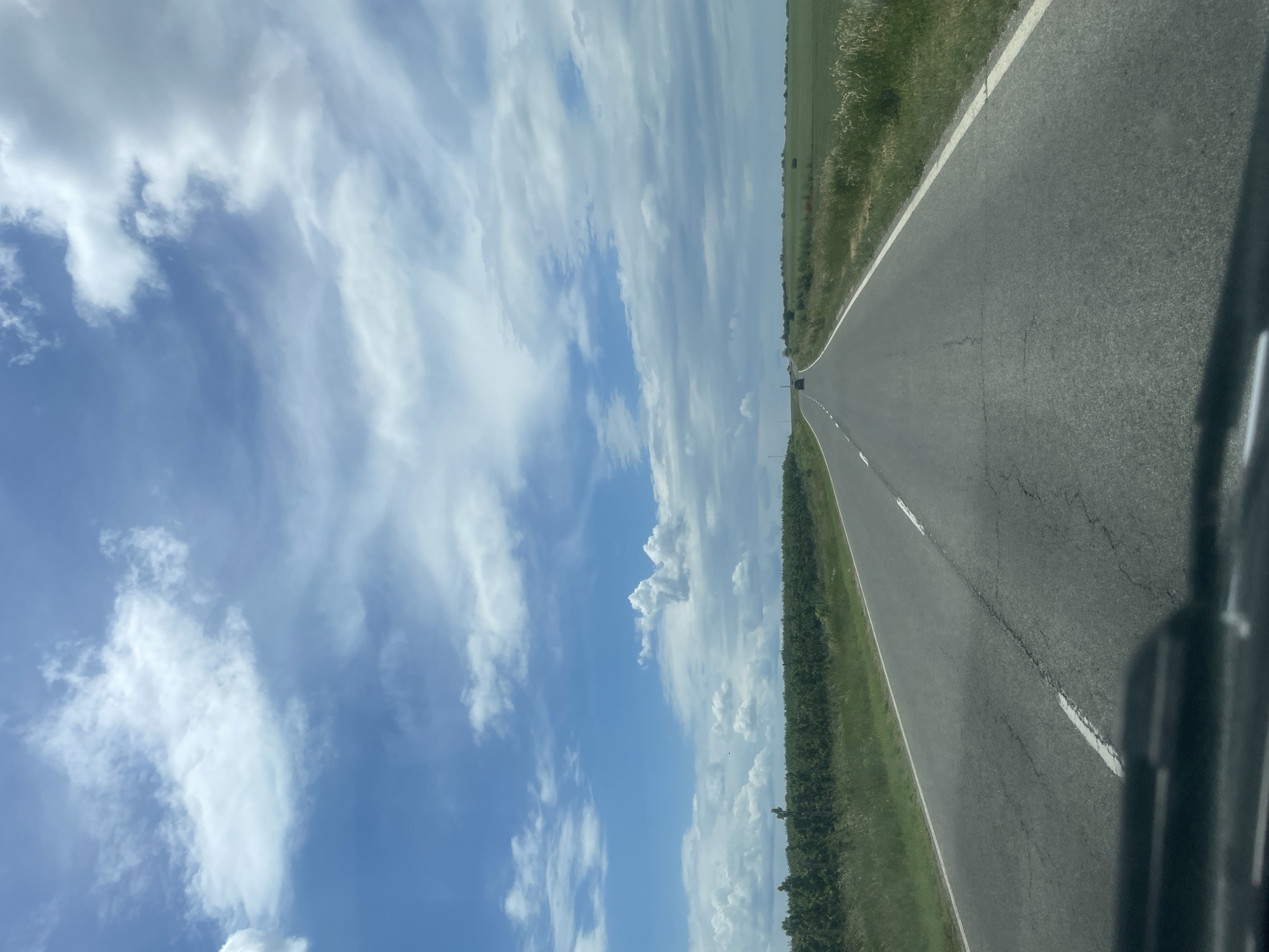 A slightly tilted view from inside a vehicle, capturing a straight, asphalt road stretching into the distance. The road is flanked by green vegetation on both sides. A blue sky with scattered white clouds dominates the upper portion of the frame. In the distance, a small, dark shape is visible on the road, presumably another vehicle. The scene may be located in Madara, Bulgaria.