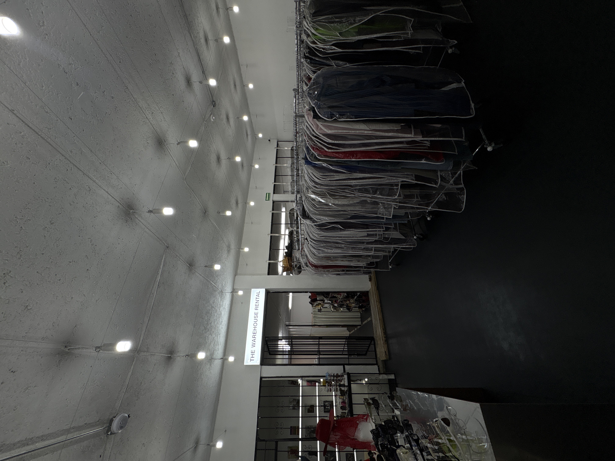 The image shows an interior view of a commercial space in Mexico City, Mexico, featuring rows of garments covered in plastic, hanging from racks. The ceiling is textured and has rows of evenly spaced lights. A sign reads "THE WAREHOUSE RENTAL." Beyond the sign, there is a view into another space with more clothing. There is no visible presence of people. A staircase with a metal railing is visible further into the interior. The right side of the image is a darkened space.