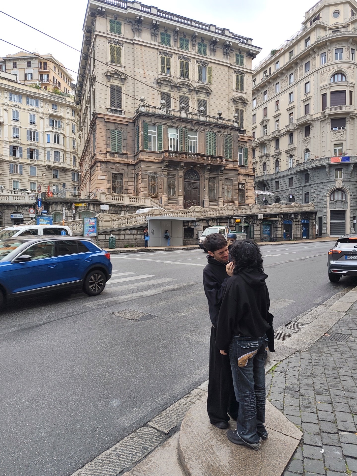 The image shows two people in Genoa, Italy, standing on a concrete platform next to a street. One person is wearing a long black coat, and the other is wearing a black hoodie and jeans with noticeable decorative details on the back pockets. They appear to be interacting intimately, possibly kissing or embracing. The street is asphalt, with a marked pedestrian crossing visible. A blue car is driving along the street. In the background, there are large, ornate buildings typical of Genoa. A gray car is parked on the side of the road behind the couple.