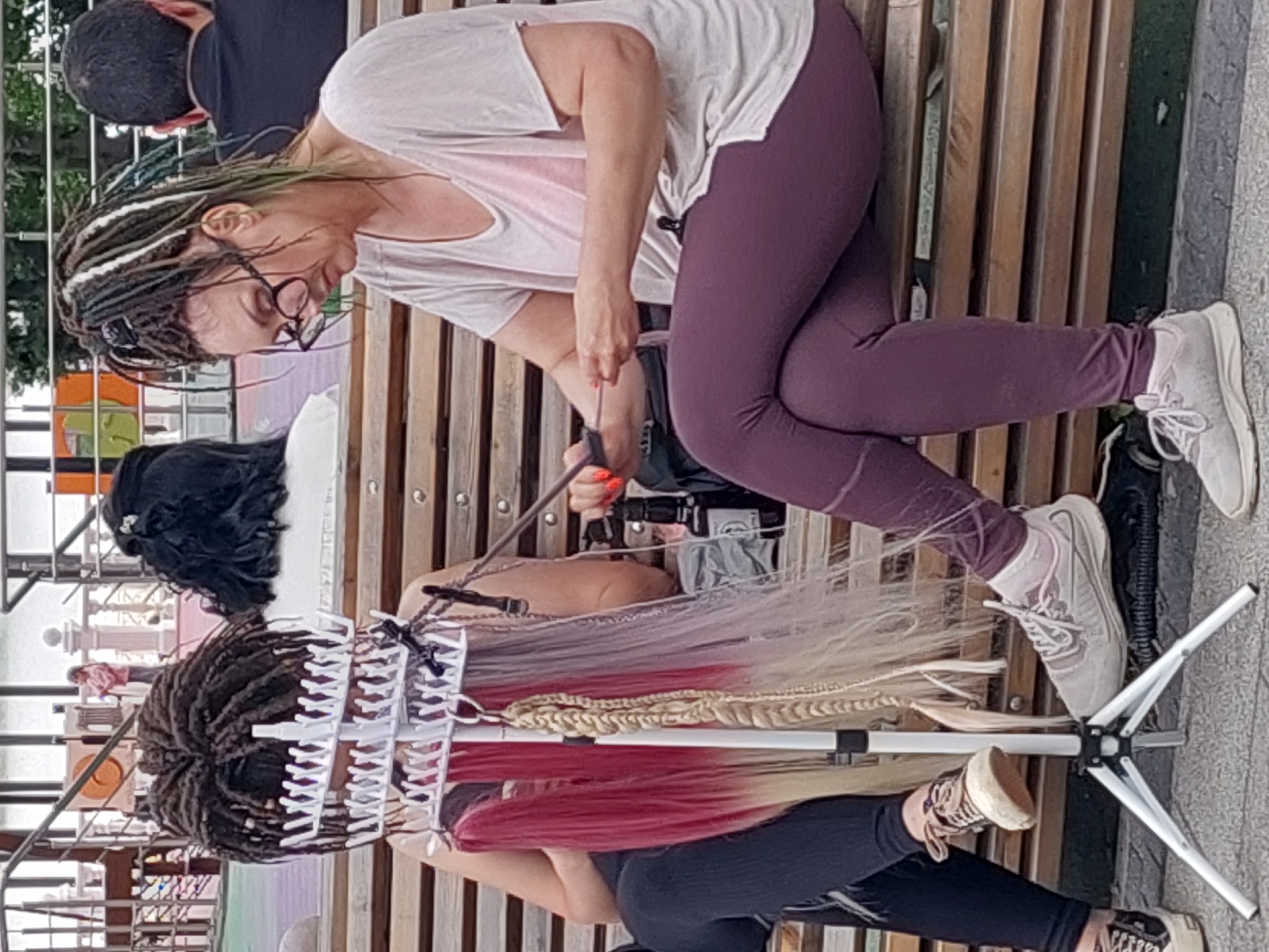An outdoor street scene in Alikonovka, Russia depicts two women sitting on a wooden bench. The woman on the left has dark dreadlocks and is partially obscured by a white rack holding various colored hair extensions. The woman on the right, wearing glasses and a light-colored t-shirt with leggings, is actively braiding hair extensions. A tripod is positioned in front of them, and a black bottle is visible on the bench. In the background, a child is seen on a playground.