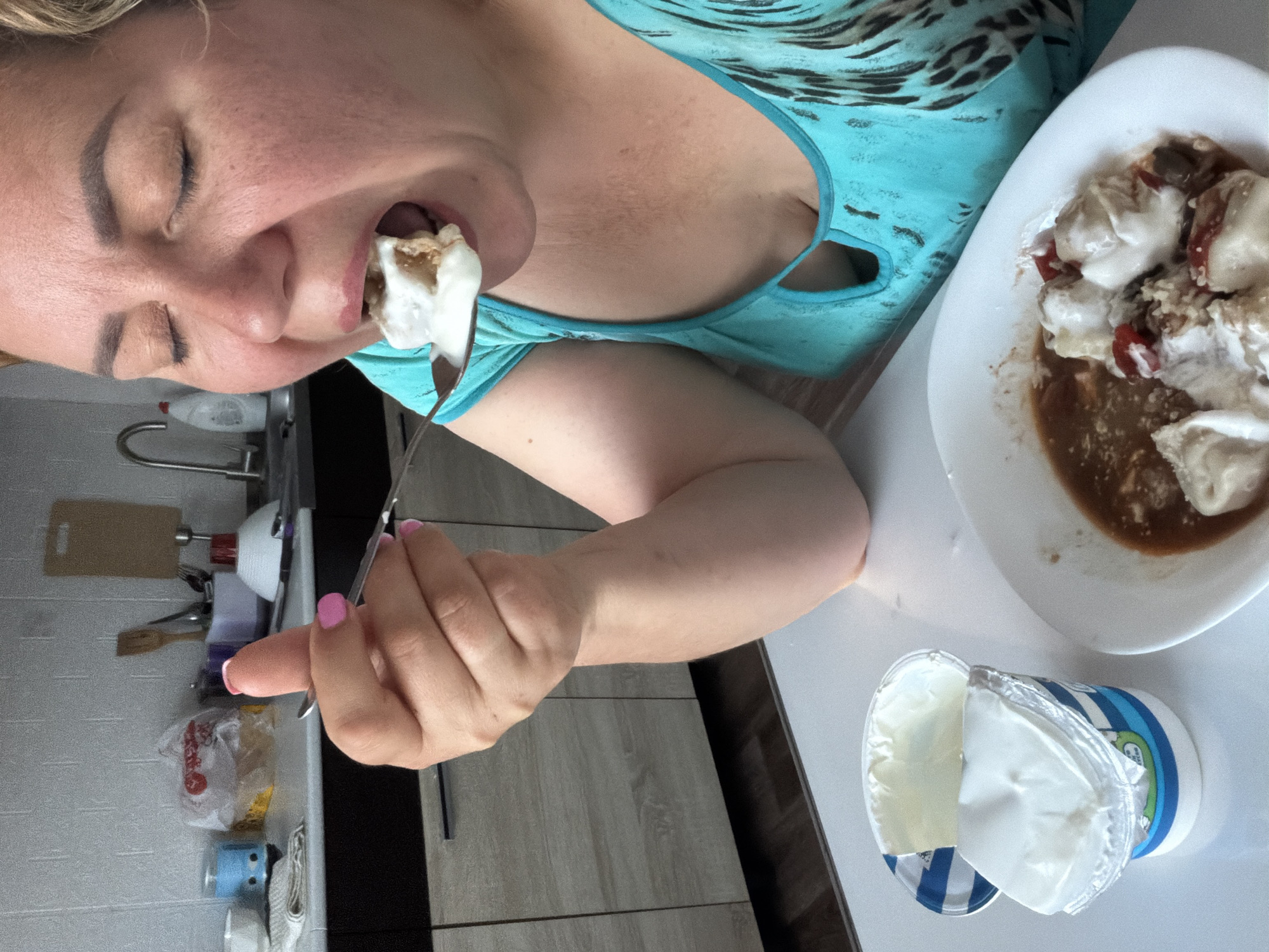 A woman with light skin and blonde hair eats a spoonful of food. The food appears to be a mixture of white yogurt-like substance, brown sauce, and red pieces, possibly tomato or pepper, on a white plate. An open container of what appears to be white yogurt sits on the table next to the plate. The woman is wearing a turquoise and black tank top. Her eyes are closed as she eats. Her fingernails are painted pink. The scene appears to be set in a kitchen, evidenced by the counter, cabinet, sink, and utensils in the background. The city is Leningradskaya, Russia.
