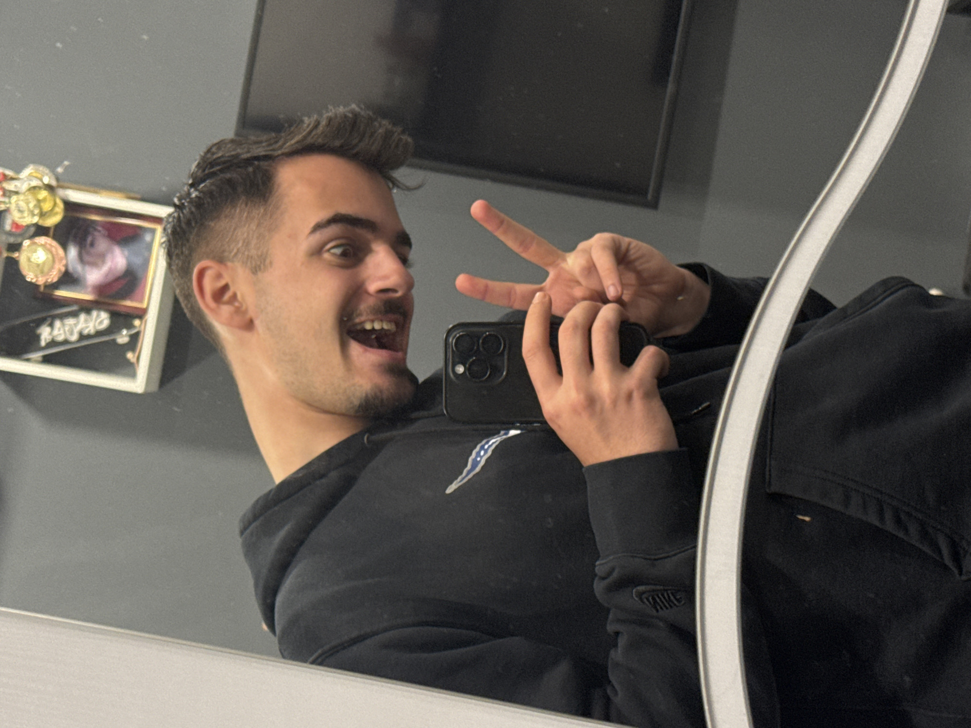 A medium shot, taken indoors, depicts a person from the chest up. The individual, appearing to be male, is angled to the left with the head turned toward the camera. The face is expressive, with the mouth open and eyes wide. The person is holding a smartphone in their left hand, presumably taking a selfie, and making a V-sign with their right hand. The subject is wearing a black hooded sweatshirt with a prominent Nike logo on the left sleeve.
The background includes a gray wall with a black-framed picture hanging on the left side. There is also what appears to be a framed photograph. The scene is shot in what appears to be a mirror, and the location in the city of Sofia, Bulgaria, cannot be determined from the image.