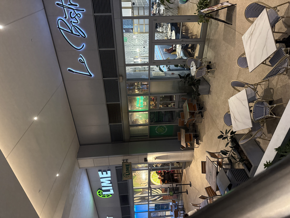 The image shows the exterior of two restaurants, "Le Bistro" and "Lime," at night. "Le Bistro" has a cursive, illuminated sign above its glass-fronted entrance. Patrons are visible inside. Outdoor seating is arranged with tables and blue and white chairs. Further down, "Lime" is visible, with its green and white sign. The restaurant also has a glass facade. Tables and chairs are set up outside.