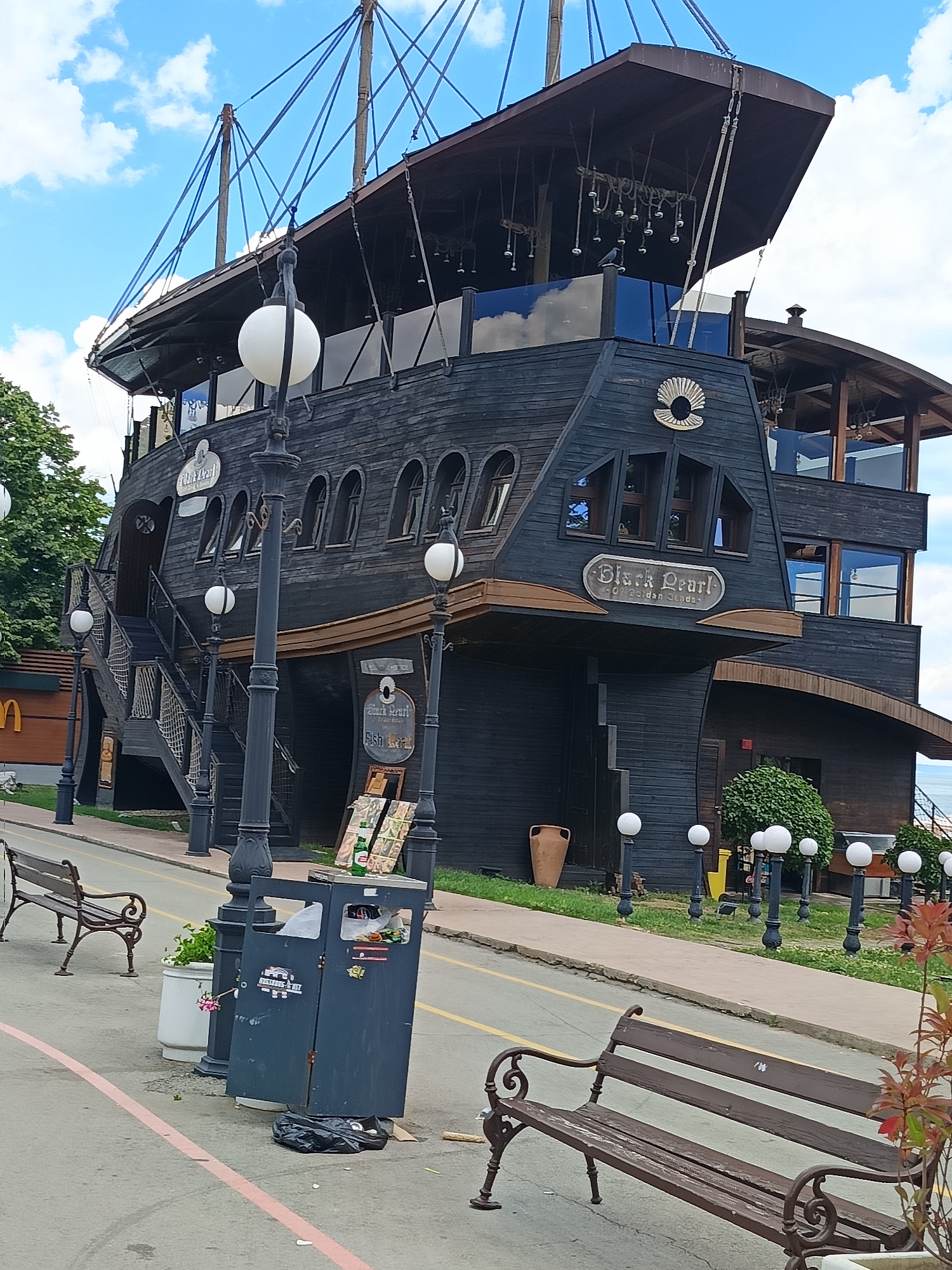 The image shows a building shaped like a pirate ship, named "Black Pearl," located in Varna, Bulgaria. The ship-like building is predominantly black with wooden accents. It has multiple levels, with windows and balconies visible. A large sign on the building reads "Black Pearl Fish Restaurant." The building appears to be situated near a waterfront, with the sea visible in the background. In the foreground, there is a sidewalk with several streetlights, benches, and a trash can. The sky is blue with some white clouds. The overall scene suggests an outdoor dining or entertainment venue with a nautical theme.