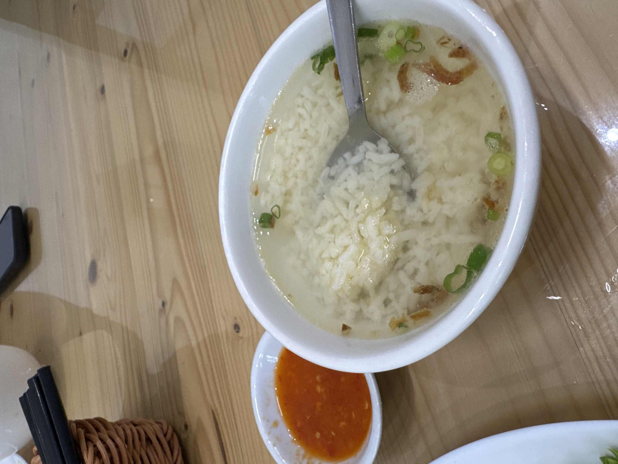 The image shows a close-up of a bowl of rice porridge with chopped green onions and some fried shallots, served with a small dish of chili sauce on a wooden table. A metal spoon is partially submerged in the porridge. In the background, there is a brown woven basket with black chopsticks and a blurred white bowl. The scene is likely in a casual dining setting. There are no people or animals visible in the image. The image does not provide any specific location information within Kuala Lumpur, Malaysia.