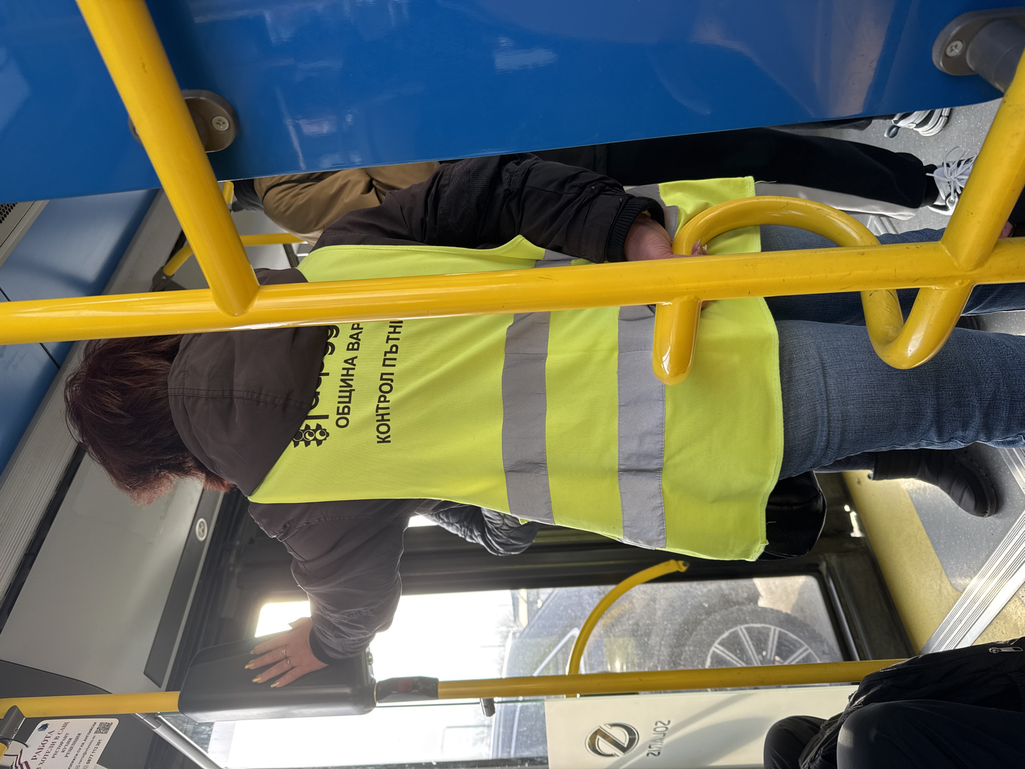 A woman wearing a yellow vest marked "Община Варна Контрол Пътни" (Municipality of Varna, Road Traffic Control) is standing in what appears to be a bus. The interior of the bus is visible, with yellow handrails and blue overhead structure. There are other people on the bus. A window is visible in the background. There is an ad with phone number 0877 113301.