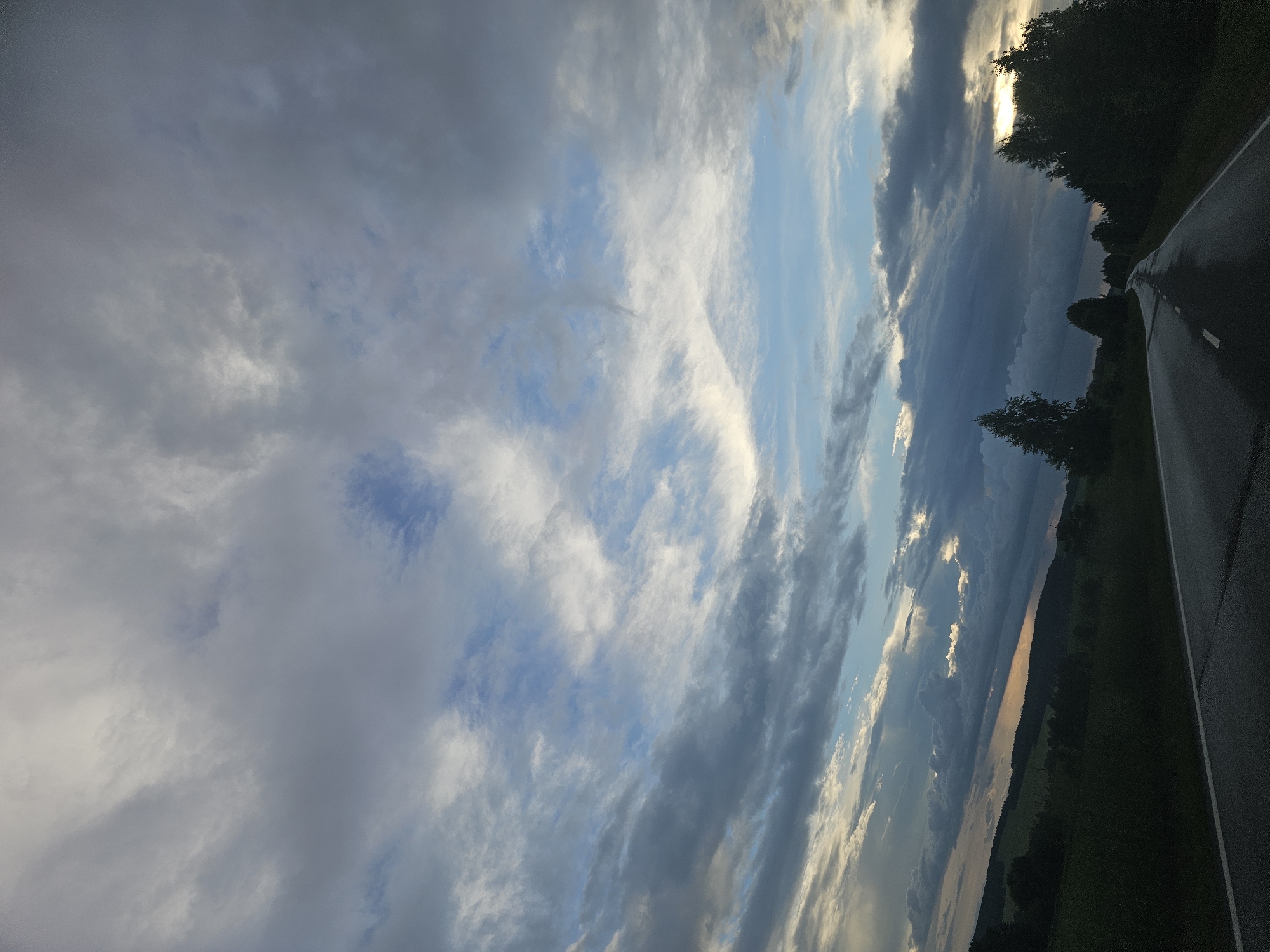 The image depicts a rural road curving through a landscape under a dramatic sky. The road, asphalt with a white dashed line, stretches into the distance, flanked by dark green vegetation and trees. The sky is a prominent feature, filled with a dynamic mix of dark, heavy clouds and lighter, wispy ones, with patches of blue sky visible in between. Rays of sunlight break through the clouds in some areas, creating shafts of light. The overall impression is of a moody, possibly pre-storm or post-storm atmosphere. There are no people or specific structures identifiable beyond the road and natural elements. The location is identified as Alnash, Russia.