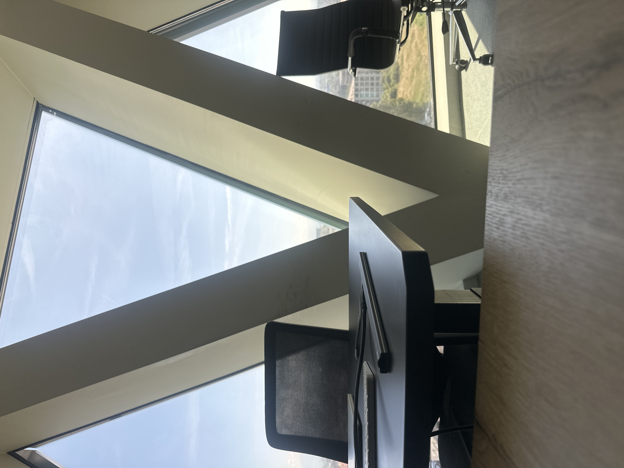 The image shows two black office chairs in front of a triangular window overlooking a city. In the foreground, there is a dark desk with a keyboard and pens. The window frame is also visible, with sunlight streaming through. The setting appears to be a modern office space. The city visible through the window is Sofia, Bulgaria.