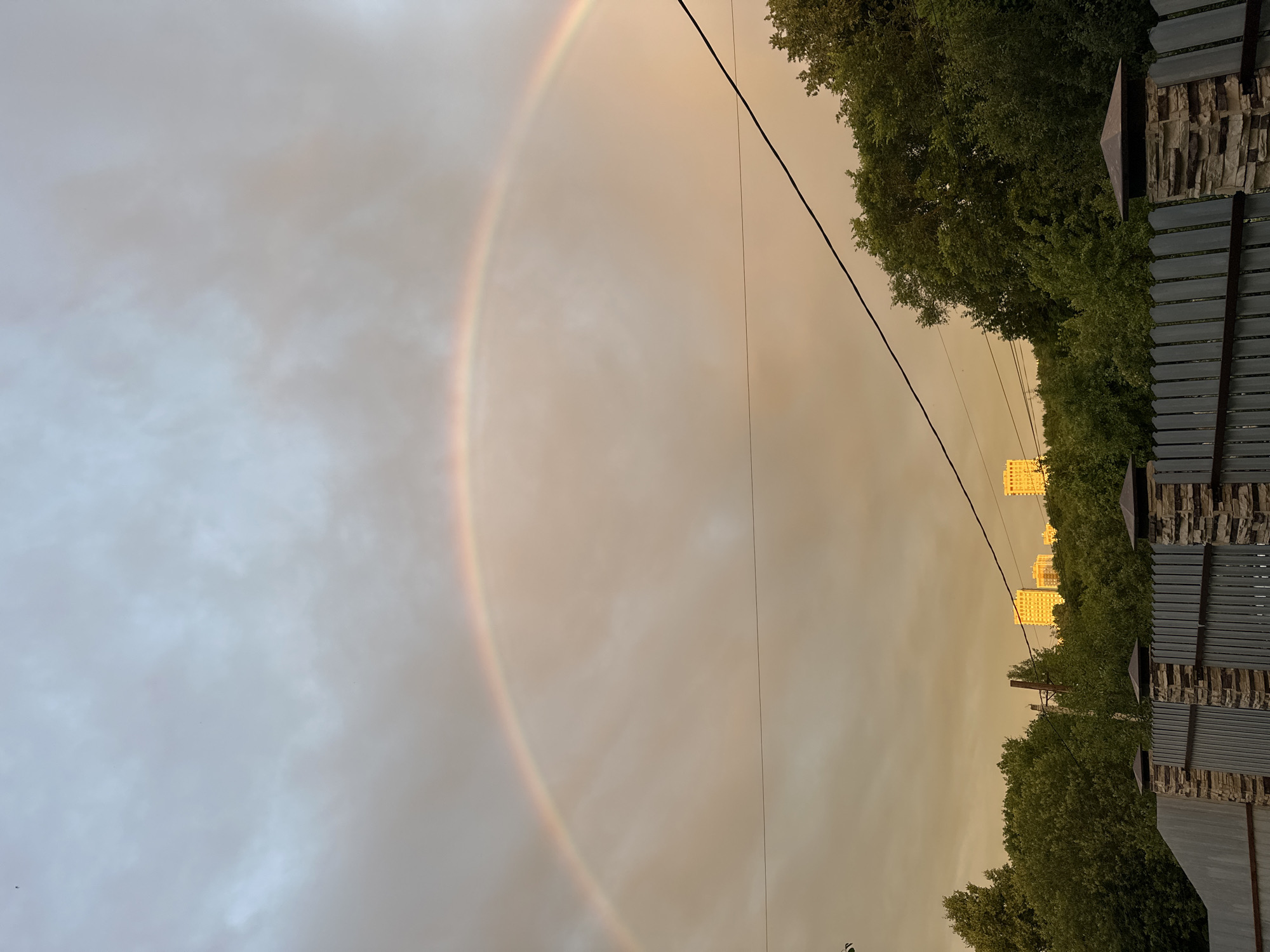 The image depicts a rainbow arcing across a cloudy sky over the city of Nizhny Novgorod, Russia. In the foreground, a fence with stone-clad pillars separates the viewer from a dense line of green trees. Beyond the trees, several multi-story residential buildings are visible, illuminated by the warm light of what appears to be either sunrise or sunset. A power line cuts horizontally across the frame, dividing the sky and the landscape. The sky itself is a mix of soft grays and warm, golden tones, suggesting atmospheric conditions conducive to rainbow formation. The overall scene captures a moment of natural beauty juxtaposed with urban development and infrastructure.