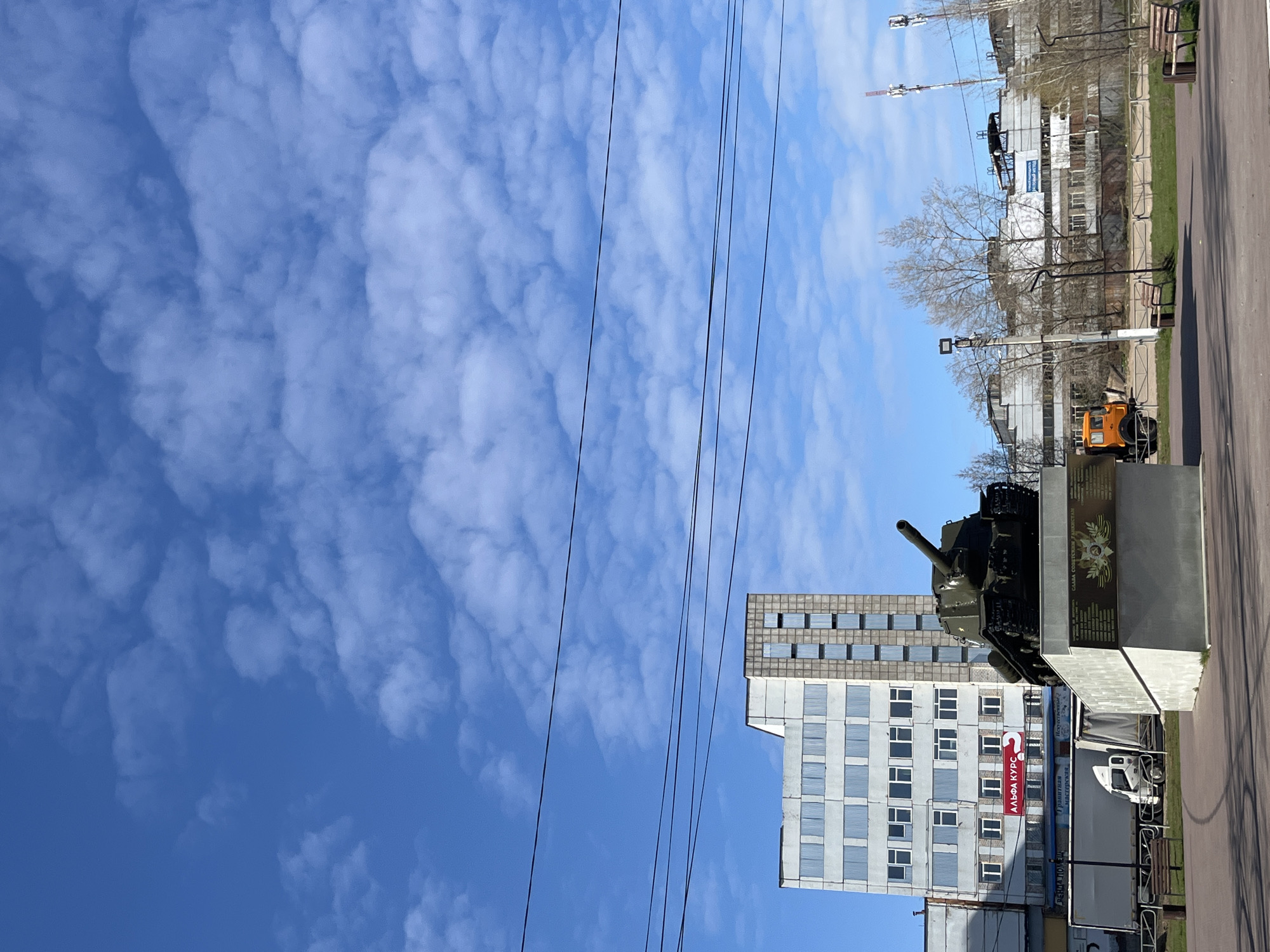 The image shows a daytime view in the city of Chaikovsky, Russia. A Soviet-era T-34 tank is displayed as a war memorial atop a concrete pedestal. In the background are buildings, including a taller structure with a grid of windows. Above, there is a blue sky with scattered clouds. Power lines are visible crossing the sky. A paved path and some greenery are also present in the foreground.