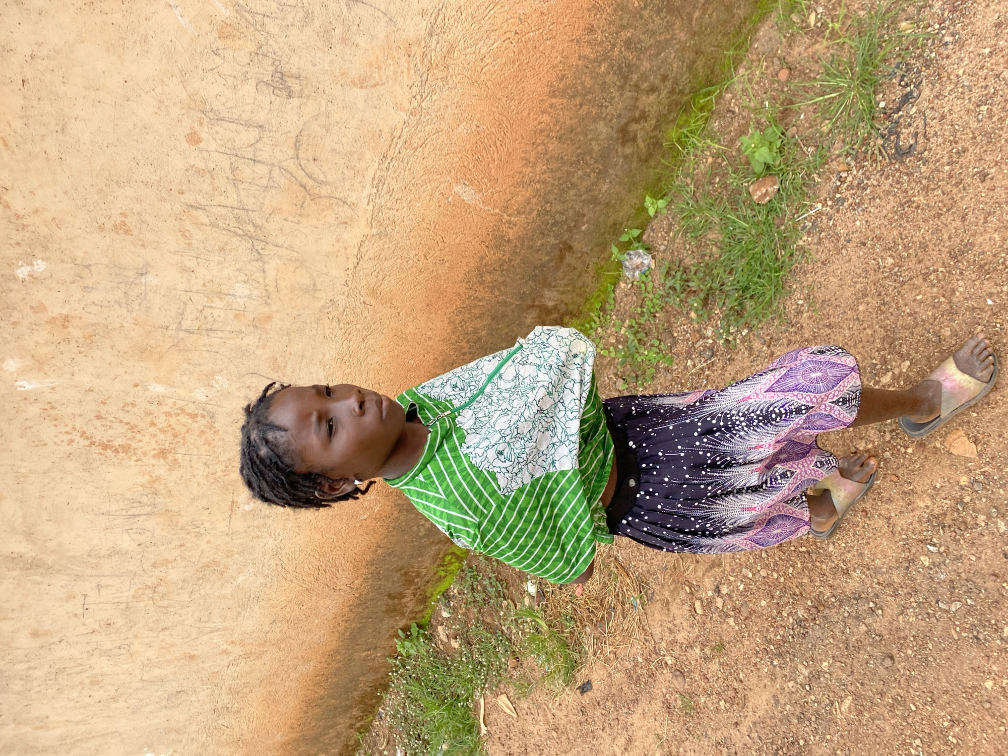 A young girl with dreadlocks stands on a dirt path in front of a textured, ochre-colored wall in Bukuru, Nigeria. She is wearing a green and white patterned shirt with a white floral design on the right side, and a dark skirt with a white and purple feathered pattern. She has on light-colored sandals. The ground is covered in reddish-brown dirt with scattered small stones and patches of green grass. The wall behind her is rough and has faint markings or graffiti on its surface. There is some trash on the ground near the grass line. The girl is looking off to her right.