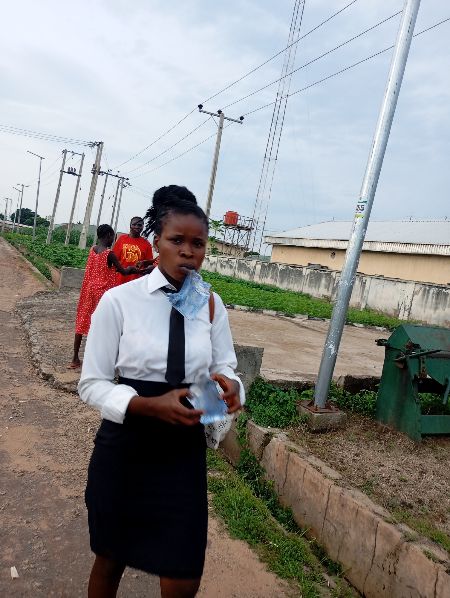 In the city of Shendam, Nigeria, a young woman in a white dress shirt and black skirt, wearing a black tie, stands on a cracked asphalt path. She is holding a partially consumed water bottle and appears to be speaking or chewing. Behind her, two other individuals, a woman in a red dress and a person in an orange t-shirt, are visible further down the path. The background features a concrete wall, lush green vegetation, utility poles with electrical wires, and buildings. A metal pole, likely a street light or utility pole, stands prominently to the right of the frame, with a green metal object, possibly machinery or a container, situated next to it. The sky is overcast.