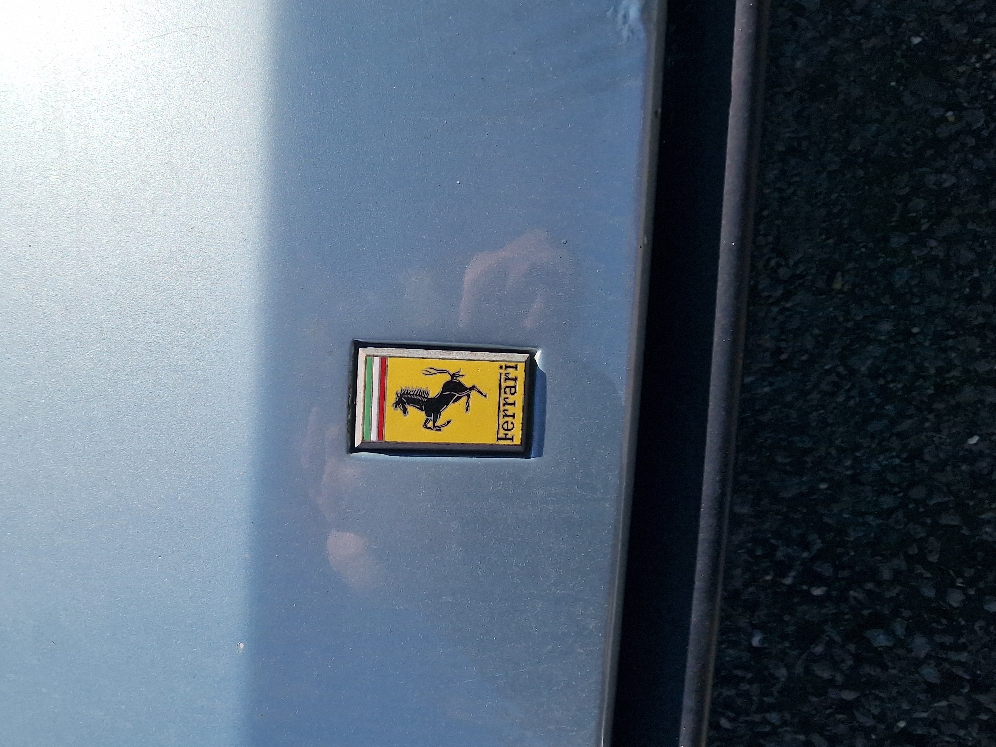 The image presents a close-up, eye-level view of a light blue or silver-grey car body panel, likely a door or fender, illuminated by bright daylight. A rectangular Ferrari shield emblem is centrally placed on the car's surface.
The emblem features the iconic black "Cavallino Rampante" (prancing horse) on a vivid yellow background. To the left of the horse, three vertical stripes in green, white, and red are visible, representing the Italian flag. The brand name "Ferrari" is written vertically in black capital letters along the right side of the yellow background. The emblem has a dark, possibly black, border.
The car's paintwork appears slightly textured or dusty, with a bright reflection of light, likely sunlight, on the upper left portion of the panel. To the right of the car panel, a dark vertical line or trim separates it from an out-of-focus background of dark asphalt or gravel, indicating an outdoor setting. No people are visible, and no specific activity is taking place. The scene suggests a clear, sunny day. The only visible text is "Ferrari" on the emblem.