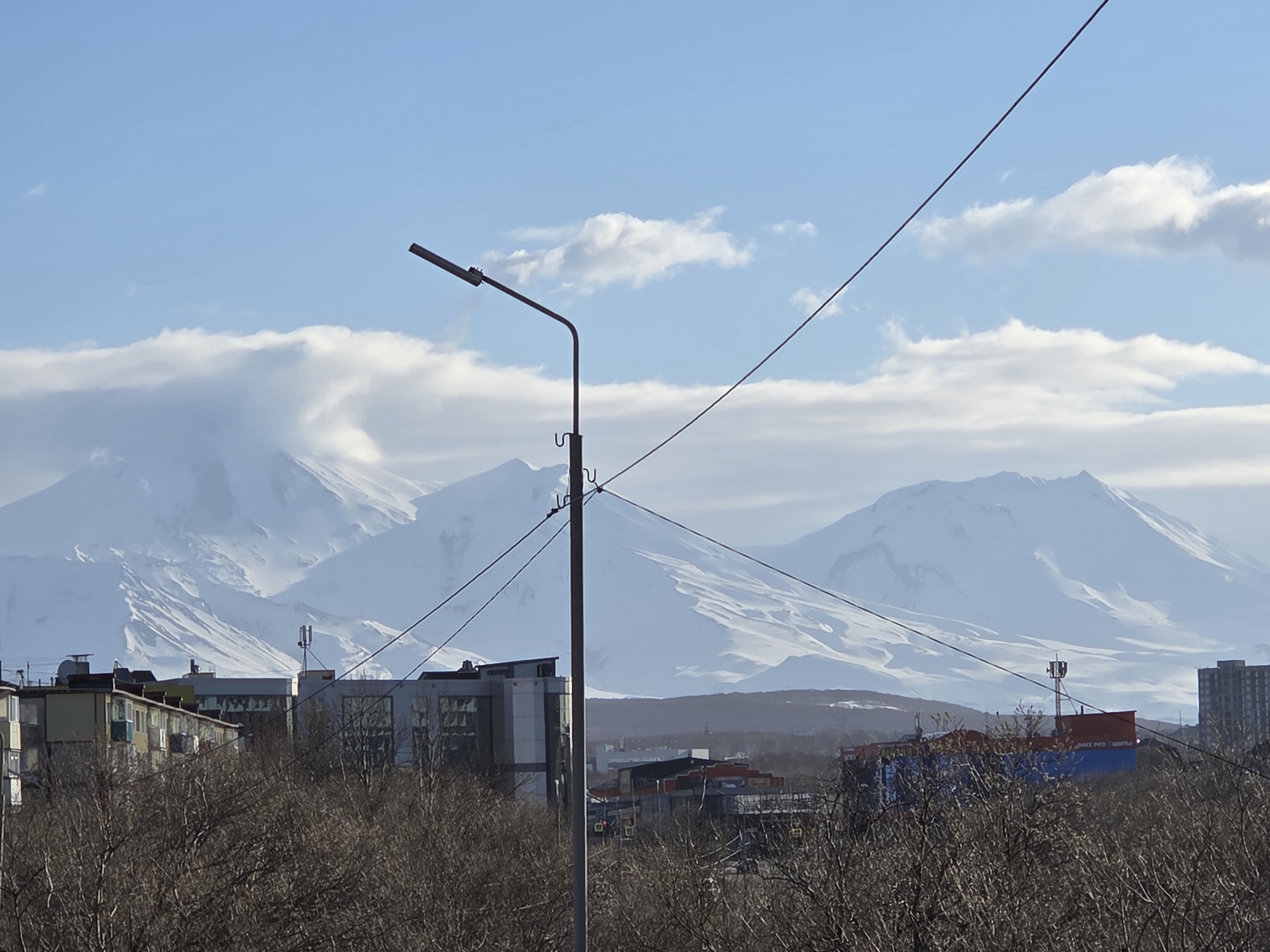 The image shows a cityscape in Unknown, Unknown, featuring a prominent mountain range in the background covered with snow and partially obscured by clouds. In the foreground, there are buildings, trees with bare branches, and a streetlight pole. Power lines cut across the sky. The sky is blue with scattered white clouds.