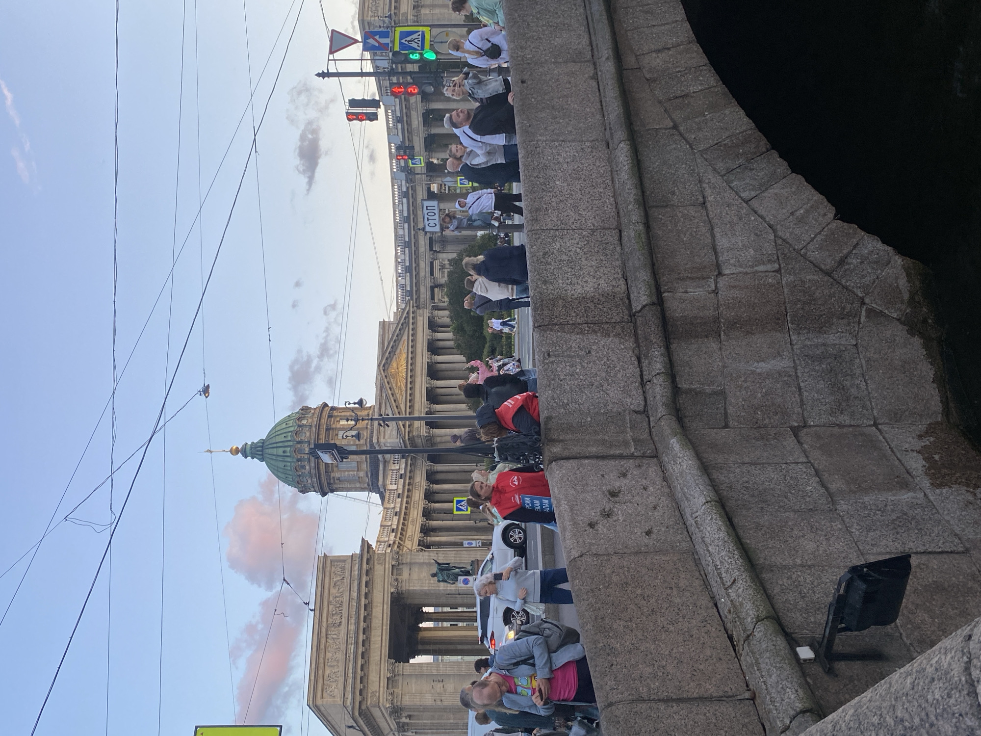 The image depicts a street scene in Saint Petersburg, Russia, likely near Kazan Cathedral, identified by the presence of the cathedral's green dome and colonnades. In the foreground, a stone bridge with a rough-hewn texture spans across a dark body of water, though the water itself is not clearly visible. A black light fixture is mounted on the side of the bridge. A crowd of people are gathered on and around the bridge, some looking towards the cathedral or the street. Traffic lights, street signs, and overhead wires are visible in the background, indicating an urban environment. A white car is parked on the street. The sky is a pale blue with some wispy pink clouds, suggesting it is either sunrise or sunset. The overall atmosphere is that of a busy public space.