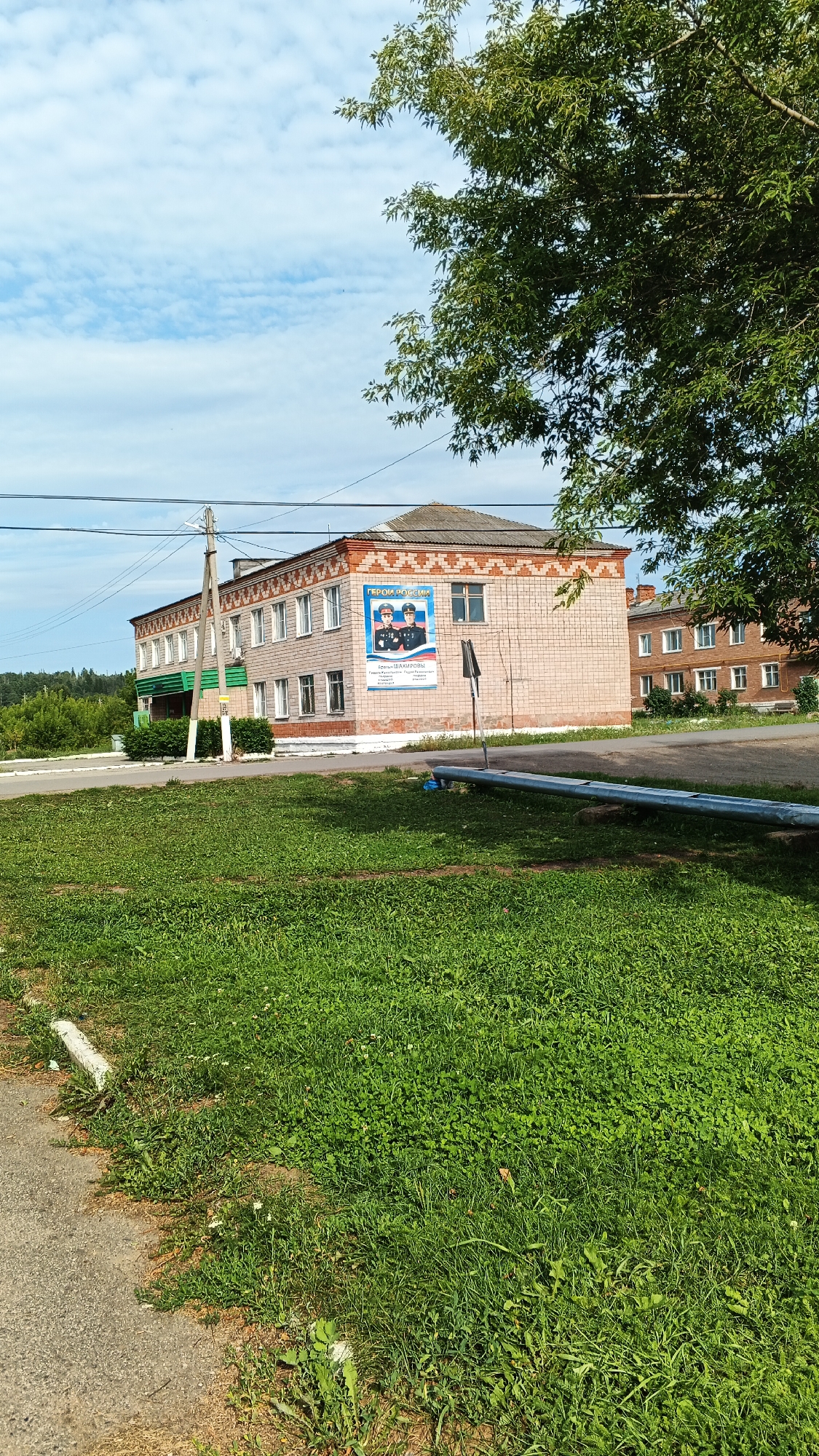 The image shows a two-story brick building with a gray roof, located in the city of Podgornoye, Russia. The building has several windows and a poster is visible on the right side of the facade. The poster features two individuals in uniform and text in Russian. In front of the building, there is a patch of green grass and a paved area. A large tree with green leaves is partially visible in the foreground, and power lines are strung across the scene. The sky is partly cloudy with scattered white clouds. The environment suggests a rural or suburban setting.