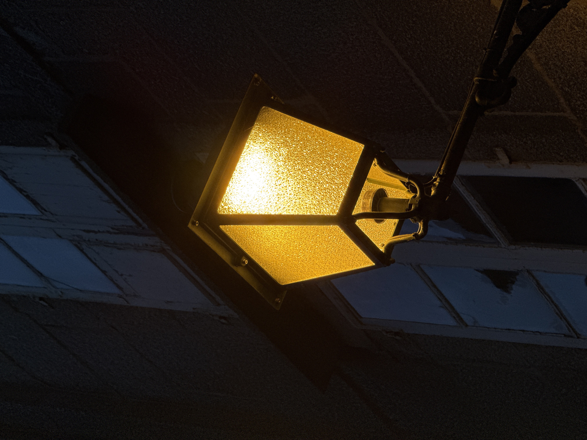 The image shows a close-up, low-angle shot of a vintage-style, yellow-lit street lamp against a dark, textured building facade. The lamp has a square, frosted glass enclosure that diffuses the warm light, and it is mounted on a black metal arm. The building appears to be constructed of stone or concrete and has several window frames visible, some of which show cracked or broken glass. The overall atmosphere is dark and moody, suggesting it is nighttime. The architectural style and the presence of a stone building are consistent with the historic city of Santiago de Compostela, Spain, though a specific location is not identifiable from the image alone. There are no people or animals present.