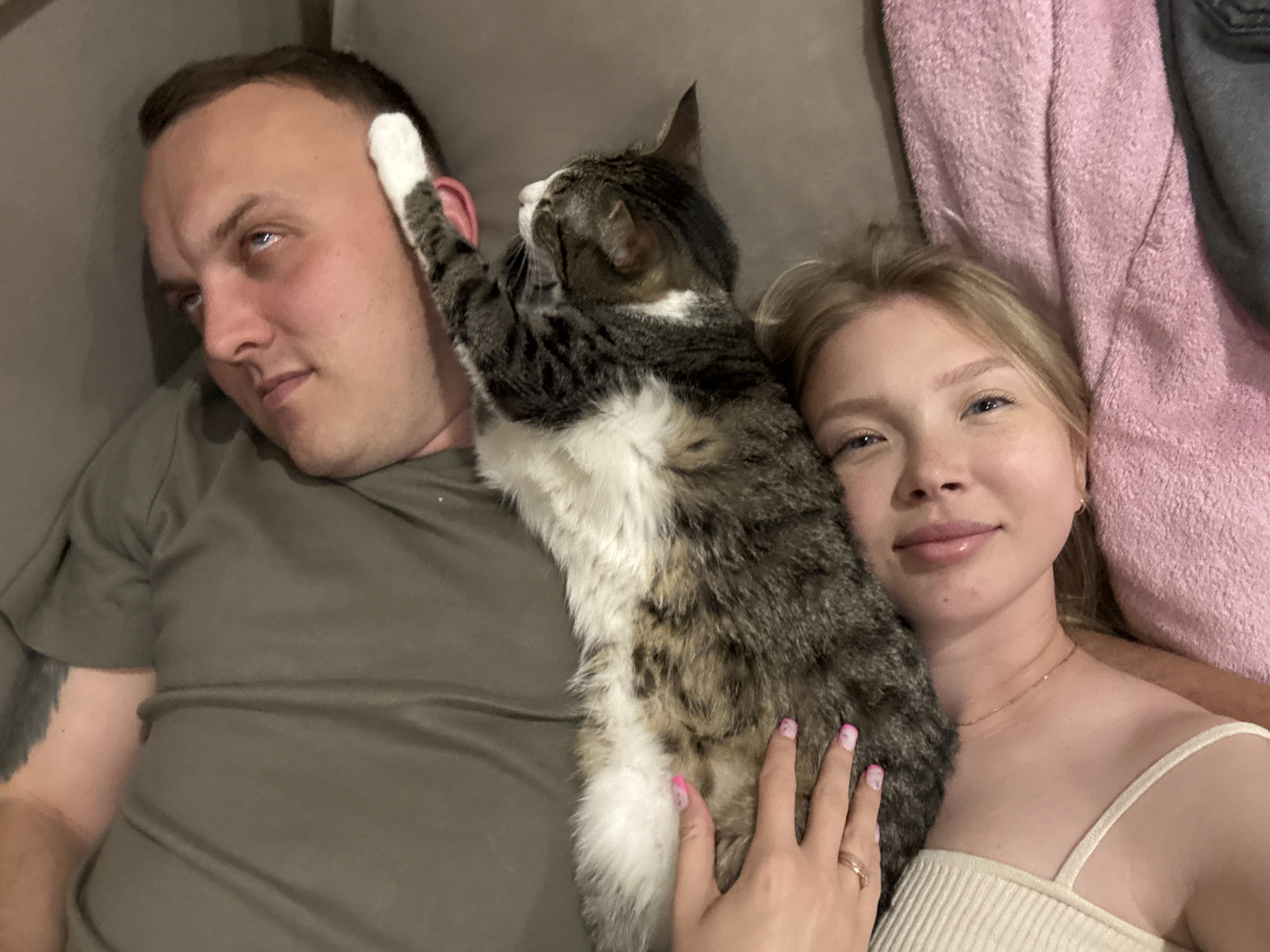 The image captures a close-up, overhead view of a couple and their cat lying down, likely on a couch or bed. The man, on the left, is wearing a dark green t-shirt and has his arm around the woman. He is looking upwards and to the left, with his eyes partially closed, giving a relaxed or perhaps playful expression. A tabby cat, positioned vertically between the couple, is playfully interacting with the man, with one of its paws raised to his head. The cat has grey, white, and black fur. The woman, on the right, is wearing a light-colored, ribbed tank top and has blonde hair. She is smiling at the camera, and her left hand, with painted nails and a ring, rests on the cat's body. A pink, fuzzy blanket is visible behind the couple, and a portion of dark gray fabric, possibly clothing or another blanket, is also present. The lighting is soft and appears to be from an indoor source.
The image is not geotagged and contains no discernible landmarks or visual cues that would allow for precise identification of the location within the city of Chaikovsky, Russia. The setting is generic and domestic.