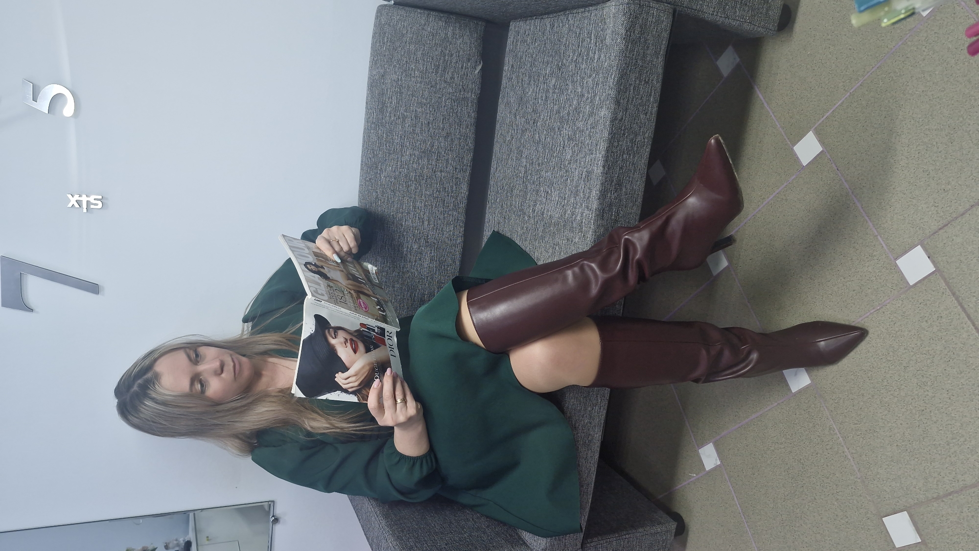 A woman is seated comfortably on a grey upholstered bench or couch, engrossed in reading a magazine in an indoor setting in Otchor, Russia. She has long, light brown or blonde hair and is wearing a dark green long-sleeved dress or tunic, paired with knee-high or thigh-high dark brown/burgundy boots. Her left hand holds the open magazine, while her right hand rests on it. The magazine displays the word "DIOR" prominently on its cover or an inside page.
The background consists of a plain light-colored wall, adorned with large, metallic numbers "5" and "7", and the word "SIX" between them, arranged vertically. The floor is covered with large, light grey or beige tiles, accented by a pattern of smaller white squares and light purple grout lines. A partial reflection, possibly from a window or another reflective surface, is visible on the lower left of the wall. The woman appears relaxed and focused on her reading in what seems to be a waiting area, lounge, or office environment. The lighting is bright and even, suggesting an indoor setting with artificial light or bright natural light.