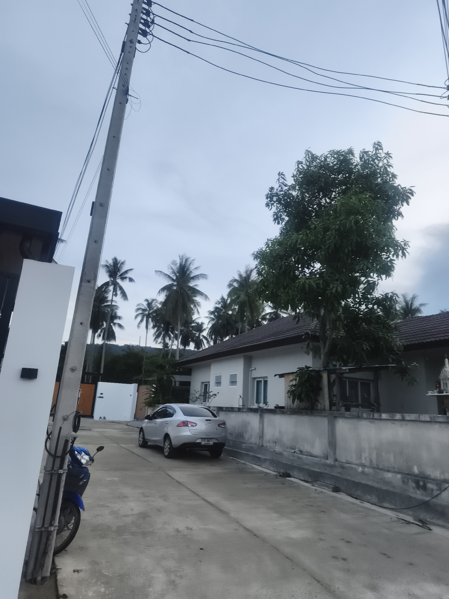 Exterior shot in Baan Mae Nam, Thailand. A concrete street leads away from the viewer, flanked by buildings and a concrete wall. A silver car is parked along the street, its rear facing the camera. A blue motorbike is parked on the left side of the street. Power lines crisscross above the scene. Palm trees are visible in the distance. The sky is overcast.