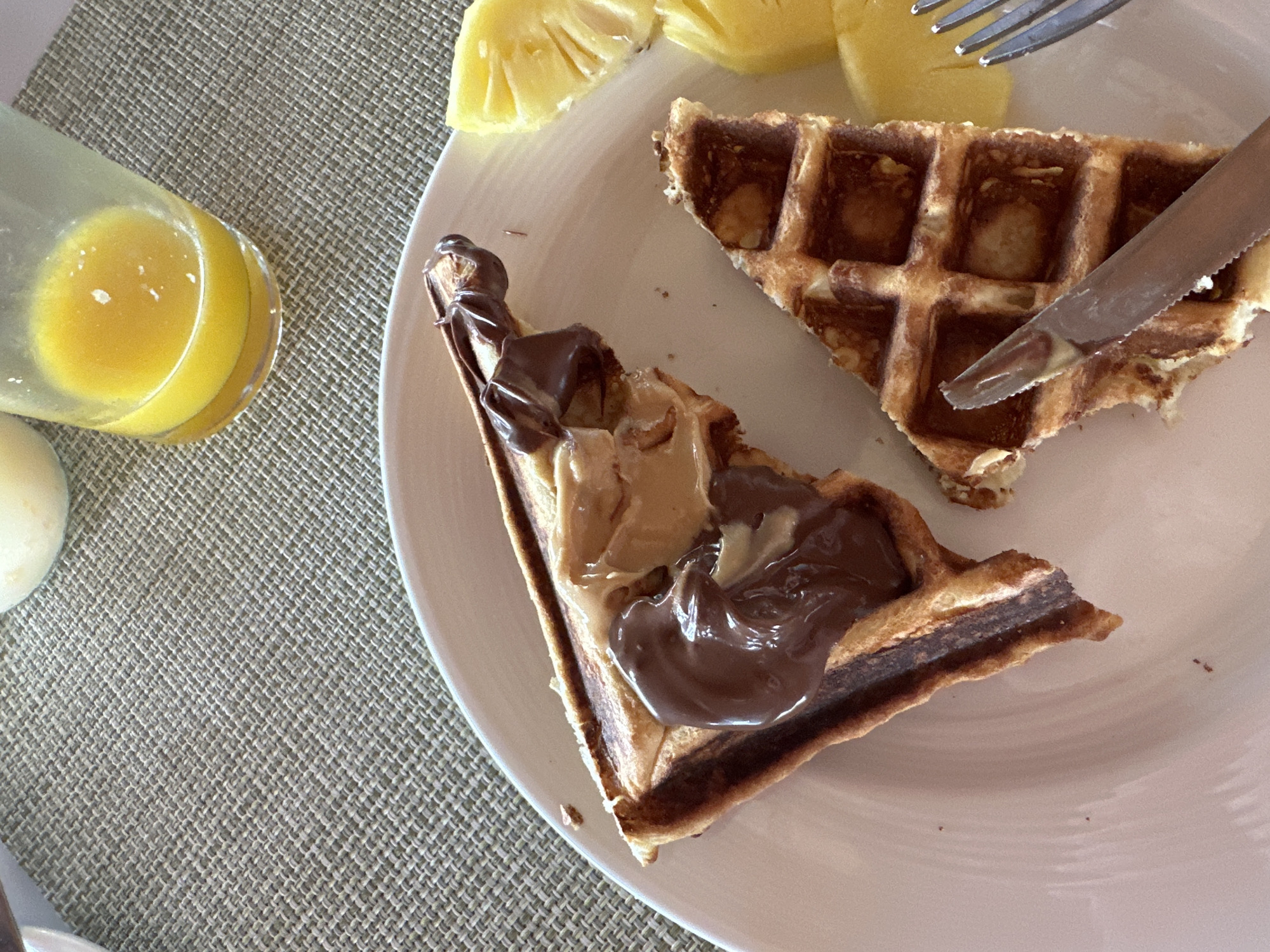 The image shows a close-up of a white plate with breakfast items. On the plate are two triangular pieces of waffle, one topped with melted chocolate and peanut butter, and the other with a knife partially obscuring it. Slices of pineapple are also on the plate. In the background, a glass of orange juice and a small bowl are visible on a textured placemat. The setting appears to be outdoors, possibly at a resort, given the presence of tropical fruit and the overall ambiance. The location is specified as Halaveli, Maldives.
