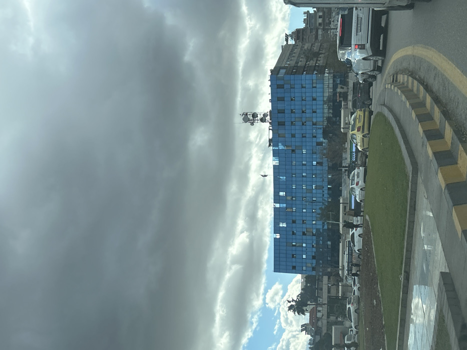 An overhead-level view depicts a busy urban street scene in Al-Muhajerin Municipality, Syria, under a predominantly overcast sky with grey clouds, interspersed with patches of lighter clouds and blue sky. The focal point is a large, multi-story building with a facade composed entirely of reflective blue glass panels, featuring numerous square windows, some appearing open or internally lit. A dark flag is flown from a pole on the roof, near the left side of the blue building, while a prominent, multi-tiered communication tower occupies the right side of its roof.
In the foreground, a paved road features a black and yellow striped curb bordering a central median of green grass, transitioning to an area of dry earth. To the far left foreground, a wet surface or puddle reflects the sky. Traffic is visible on the road, including several sedans of varying colors (white, yellow) and a white SUV positioned prominently on the right. Pedestrians are observed near the median on the left side of the street, walking or standing. Additional buildings, including one with a red roof and visible palm trees, line the background on the left, while other urban structures extend to the right of the central blue building.