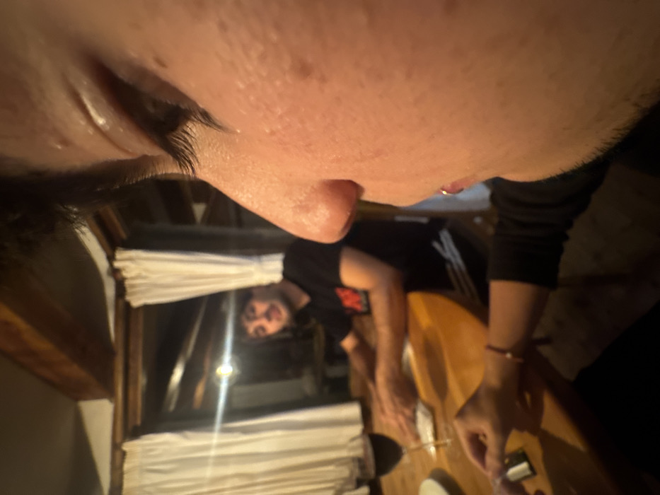 A close-up perspective from above captures a person's eye, cheek, and nose. The background reveals a man reflected in a window, wearing a black shirt and Adidas pants. He appears to be at a dining table with another individual, whose hand is visible. The table is wooden, and curtains adorn the window. The lighting is dim, giving the scene a warm, intimate feel.