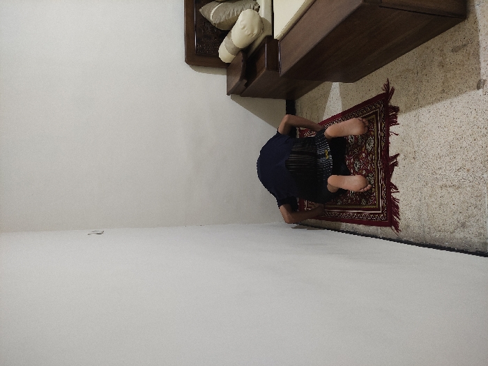 The image shows a man kneeling in prayer on a red prayer rug, his head bowed in front of a plain white wall, in a room located at coordinates (-7.828, 112.701) in Kecamatan Lawang, Indonesia. The man is wearing a dark long-sleeved shirt and dark pants. The prayer rug has a fringe border. There is a partially visible wooden bed frame with cushions to the right of the man, and a matching wooden footboard or nightstand with a dark surface in the lower right corner. The flooring is a light-colored speckled tile, and the walls are white, with a light switch visible in the upper left corner of the image. The room is otherwise empty.