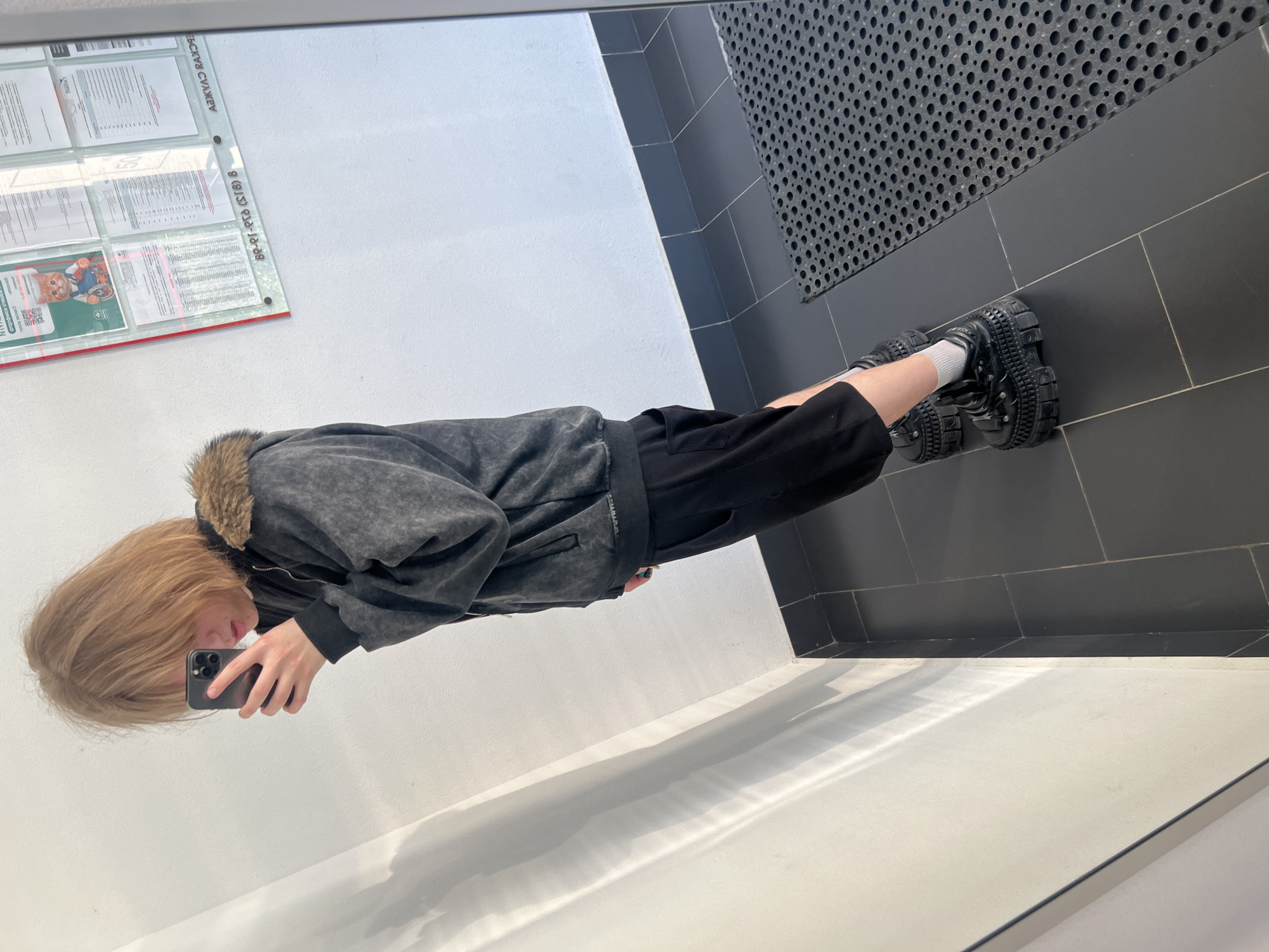 An indoor mirror selfie captures a young individual in a hallway or corridor in Sinyavino, Russia. The person, with light brown, shoulder-length hair and fair skin, is angled towards the mirror, holding a dark-colored smartphone in their right hand, seemingly taking the photograph. They are dressed in a dark grey, possibly stone-washed, zippered hoodie with a brown faux-fur trim on the hood, black shorts, white ankle socks, and black, heavily-platformed shoes with textured soles and uppers. A glimpse of blue/green nail polish is visible on one finger.
The environment features white walls, with diagonal shadows and light reflections visible on the left side, indicating an external light source. The floor is covered in dark grey tiles with visible grout lines, leading to a dark tiled baseboard. A black rubber doormat with a repeating pattern of circular perforations is positioned on the right side of the floor. On the reflected wall to the right, a glass-covered bulletin board with a red trim displays several documents. One document prominently features a cartoon orange cat in a wheelchair, accompanied by Russian text including "ДЛЯ СЛАБОСЛЫШАЩИХ" (FOR THE HARD OF HEARING). Other documents contain dense Russian text, and reflected Russian text "АЗБУКА ЗАКОН" (Alphabet of Law) along with a partial phone number "89-91-918 (813) 8" are discernible on the board's surface.
