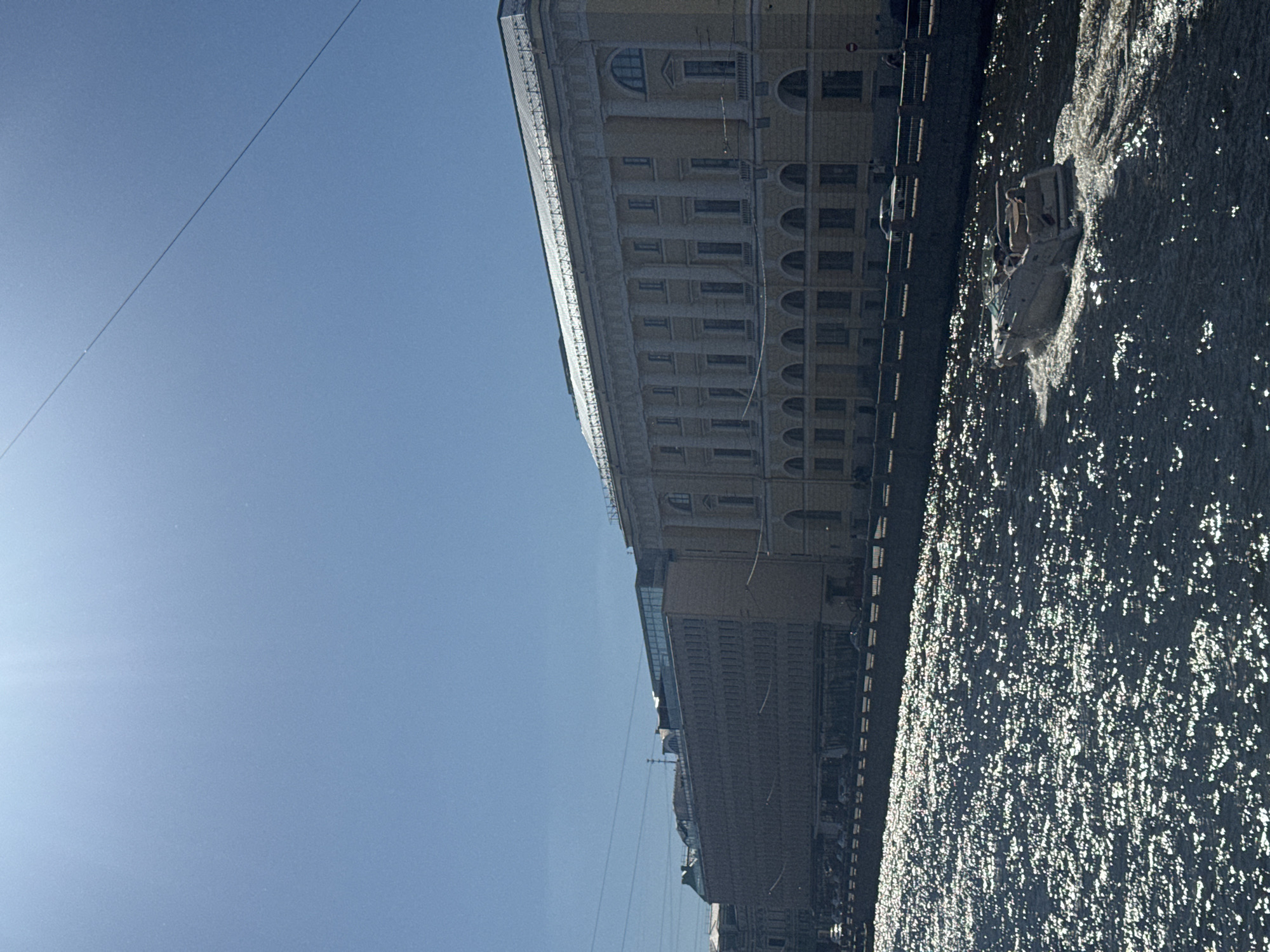 The image depicts a bright, sunny day in Saint Petersburg, Russia, with a clear blue sky. The scene focuses on a wide canal or river with a boat actively moving on its surface, creating a wake. The water is heavily dappled with sunlight, reflecting brilliantly. Along the embankment, there is a row of large, multi-story buildings with classical architectural elements, including columns and arched windows. One building on the right is pale yellow, while the building to its left is a darker brick color and appears to be a more modern addition with a glass-paneled upper section. A fence or railing lines the embankment, and there are a few cars visible on the street behind it. The overall impression is one of urban architecture meeting a sunlit waterway.
