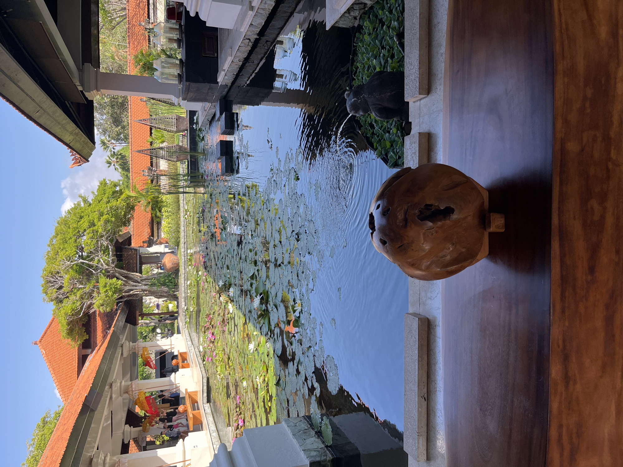 This is a medium shot, outdoor, eye-level photograph of a pond filled with lily pads and flowers in the city of Kuta Selatan, Indonesia. In the foreground, a wooden table with a polished surface is visible. On the table, a large, irregularly shaped wooden sculpture with a hollow center is placed. Beyond the sculpture, a pond with dark blue water occupies the center of the frame. Numerous green lily pads float on the surface, interspersed with white and pink lily flowers. A small, dark statue of a frog is positioned to the right of the pond, surrounded by lush green plants, and a gentle fountain is visible in the water. In the background, traditional Balinese architecture with red-tiled roofs is present, supported by white pillars. Several people, some holding colorful umbrellas, are seen walking under a covered walkway on the left side of the pond, near the building. The sky above is clear and blue. The scene conveys a serene and tropical atmosphere.