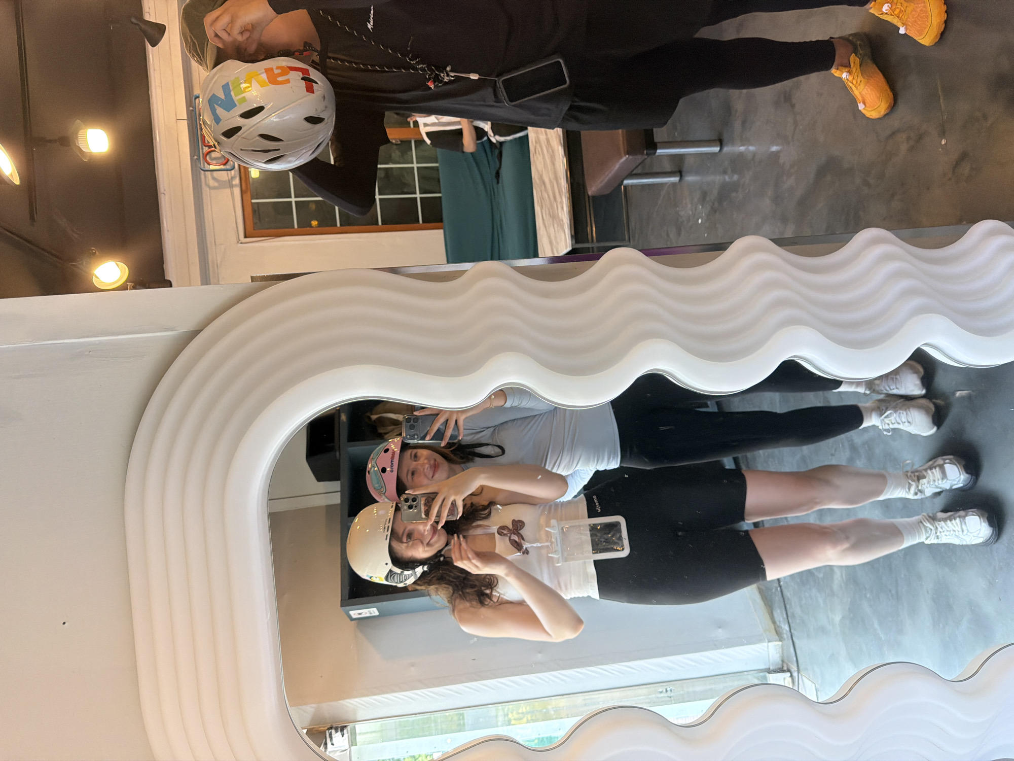 In the city of Seo-myeon, South Korea, two young women are taking a mirror selfie in what appears to be a casual indoor space. The woman on the left, with long brown hair, wears a white tank top, black shorts, white socks, and white sneakers. She has a beige helmet on her head and is holding a smartphone in her right hand, with a transparent pouch hanging from her neck. The woman on the right, with darker hair, is wearing a light blue long-sleeved shirt, black leggings, and white sneakers. She has a pink helmet on her head and is also holding a smartphone. Another individual, seen in the reflection behind them, is wearing a black shirt and shorts and has an orange helmet with colorful writing. The background includes a shelf unit, a sofa, and a table. The mirror itself has a distinctive wavy white frame.