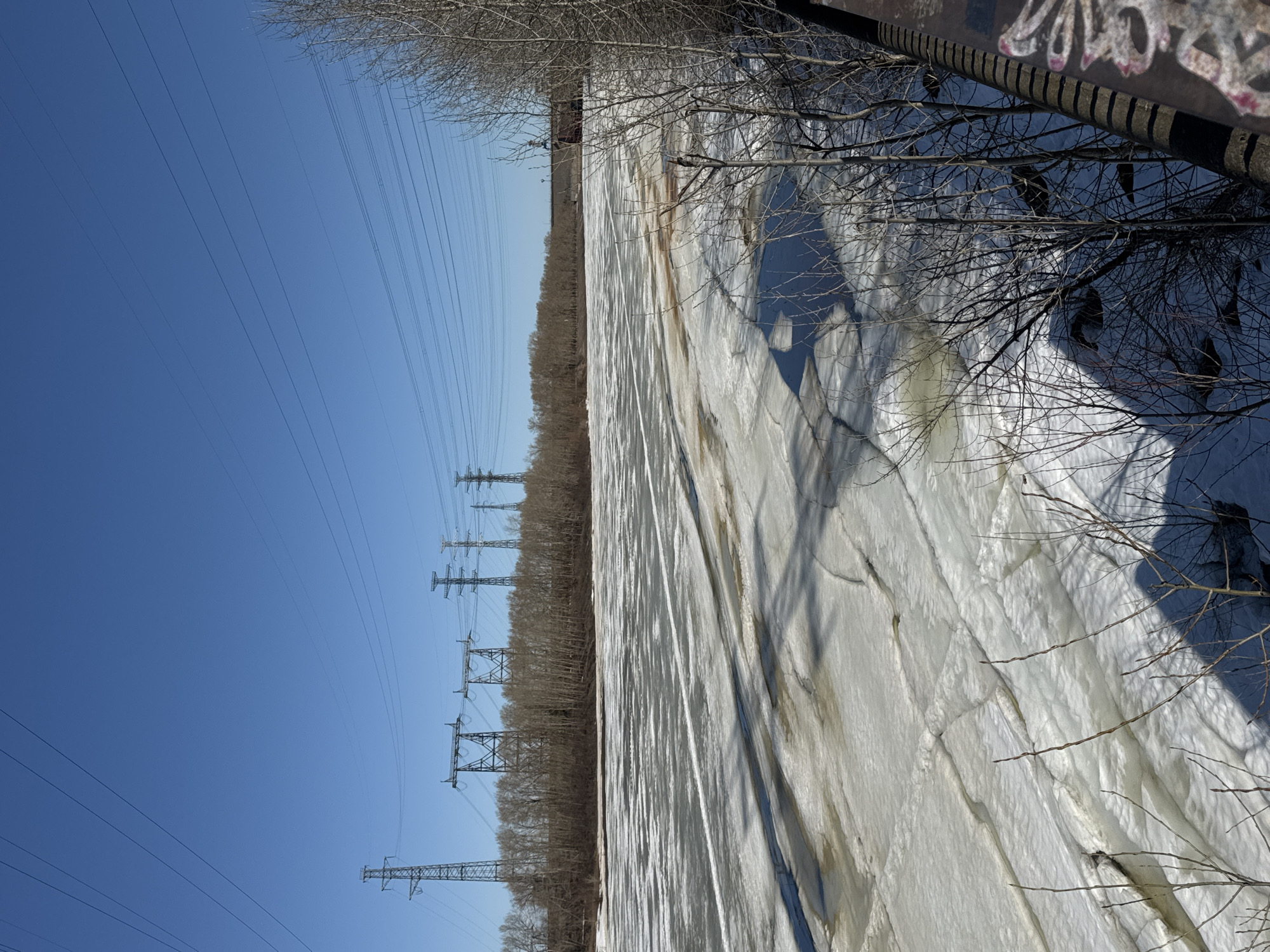 The image depicts a landscape in Chaikovsky, Russia, characterized by a field covered in what appears to be snow or ice, possibly a frozen body of water. Power lines stretch across the left side of the frame against a clear blue sky. Tall, leafless trees are visible along the middle ground and on the right, with some branches casting shadows on the icy surface. The texture of the snow/ice is rough and uneven. A structure, possibly a bridge or building with graffiti, is visible at the top right corner of the frame.