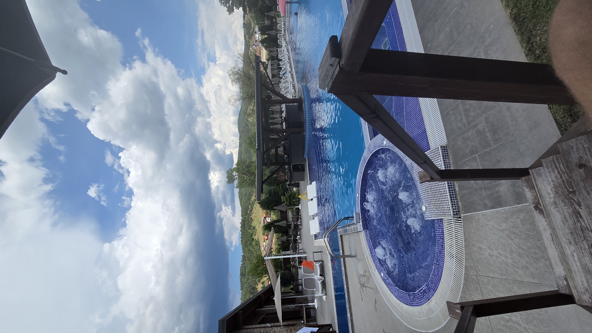 A wide-angle shot captures an outdoor swimming pool area under a cloudy sky. The pool, filled with bright blue water, is surrounded by a tiled edge and a grey tiled deck. A circular jacuzzi with bubbling water is situated in the foreground to the left. In the background, there is a covered bar area with bar stools and numerous lounge chairs lined up. To the left of the pool is a building with a stone facade and an outdoor seating area with tables and chairs. Beyond the pool area, rolling hills with scattered houses and trees are visible, under a dramatic sky with both bright blue patches and dark, heavy clouds. Wooden stairs lead down to the pool area from the foreground.