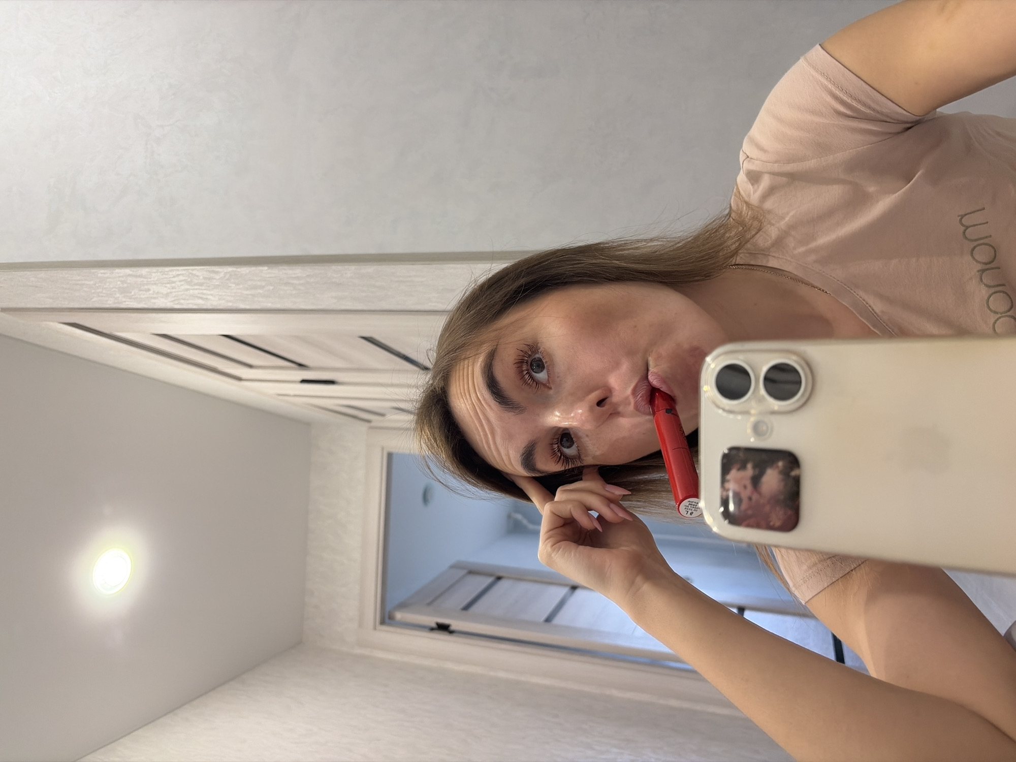 A woman is applying lipstick and taking a selfie in what appears to be a bathroom. The woman has fair skin, light brown hair, and is wearing a light pink t-shirt. She is holding an iPhone in her right hand and a red lipstick tube in her left hand, which she is applying to her lips. The bathroom has a white door with horizontal panels and a recessed ceiling light. The image does not contain sufficient information to determine the location within the city of Sarapul, Russia.