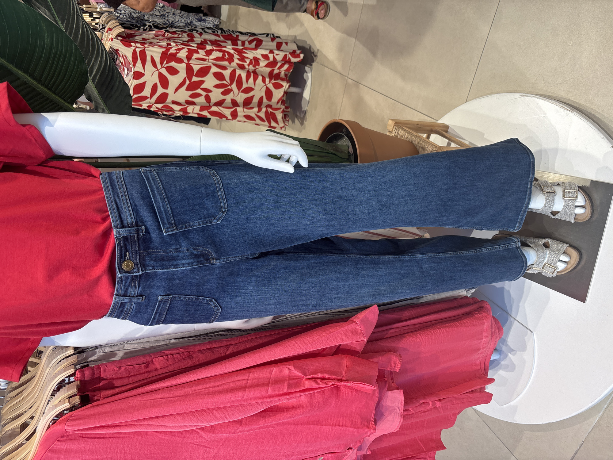 In a retail store in Orihuela, Spain, a mannequin is displayed wearing a red t-shirt and blue flared jeans. The jeans have a high waist and two patch pockets on the front. The mannequin is also wearing a pair of neutral-colored sandals with buckles. To the left of the mannequin are racks of clothing, including a bright pink long-sleeved shirt and other garments. Behind the mannequin, a potted plant is visible, and further in the background, other clothing items are displayed on racks, including a dress with a red and white floral pattern. The flooring appears to be tiled.