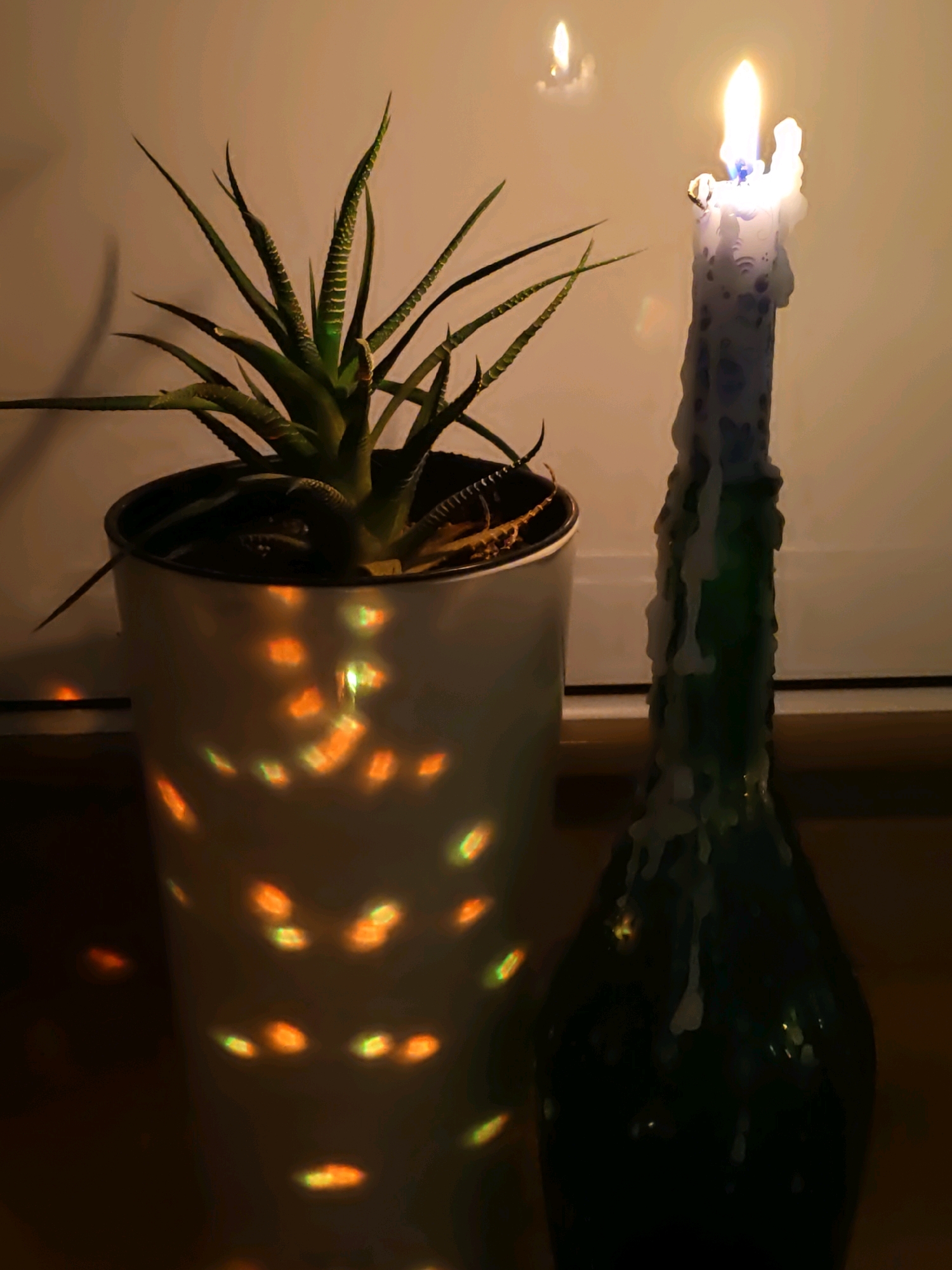 This image depicts an indoor still life scene. In the foreground, on the left, is a potted succulent plant with green, spiky leaves. The pot is white and cylindrical, and it displays colorful, blurry reflections of light on its surface, suggesting a light source with a patterned effect is nearby or behind it. To the right of the plant is a dark green glass bottle, likely a wine or liquor bottle, with a thick, white candle placed on top. The candle is lit, with a bright flame at its tip and wax dripping down its sides, creating an uneven texture. The background is a soft, muted orange-brown, possibly a wall or curtain, with a faint reflection of a light source visible in the upper right quadrant. The lighting is dim, creating a warm and somewhat intimate atmosphere. The scene is devoid of any people or identifiable urban features. Given the provided context, this scene is located in Rostov-on-Don, Russia.
