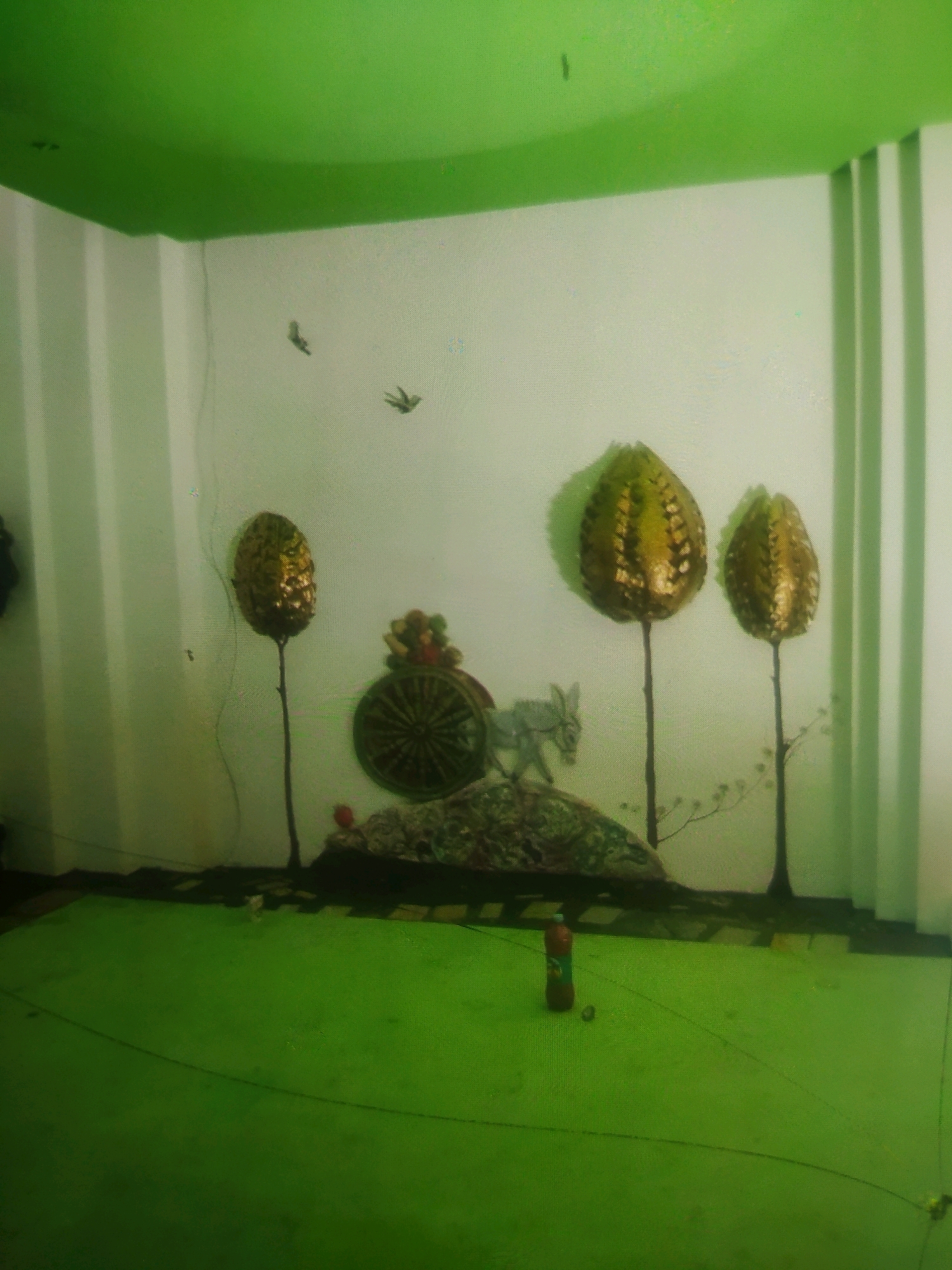 In a room with bright green ceiling and floor, a white wall features several decorative elements. To the left, a single elongated, golden, pineapple-like object stands on a thin, dark stalk. Further to the right, a cart with a circular, patterned wheel is decorated with colorful baubles. Beside the cart, a small, grey, donkey-like figurine is positioned. Three more of the golden, pineapple-like objects, also on dark stalks, are arranged on the wall. Two small, dark bird silhouettes are visible on the upper portion of the wall. The floor is covered with a bright green material, and a dark, irregular border runs along the base of the wall. On the floor, a red plastic bottle and a small dark object are present. The environment suggests an indoor setting, potentially a display area or a room decorated with a theme, located in Birlik, Uzbekistan.