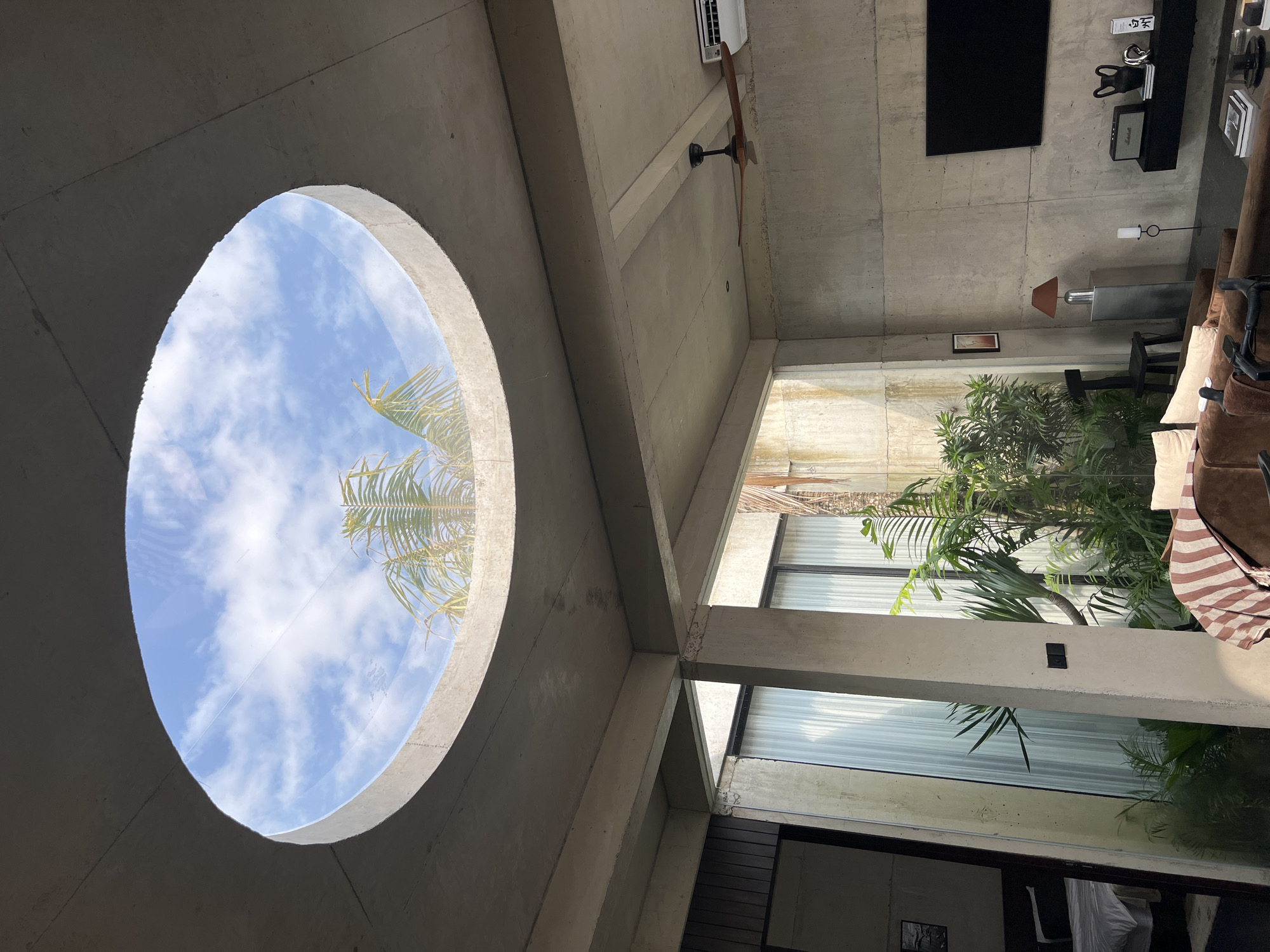 The media file presents an interior view of a modern architectural space, likely a living area or lounge, characterized by exposed concrete walls, ceiling, and structural beams. The perspective looks upwards and across the room.
A dominant feature is a large circular opening in the ceiling, serving as a skylight, offering a clear view of a bright blue sky dotted with fluffy white clouds. Lush green palm fronds are visible through this opening, extending into the sky.
The room is furnished with a large, comfortable-looking brown sofa with pillows and a striped blanket draped over it, positioned against a wall with a large window. Tropical plants are visible both outside and seemingly within the space behind glass panels, connecting the interior with nature.
On the concrete wall to the right, a large, dark flat-screen television is mounted. Below it, a long, dark shelf holds various decorative objects, including what appears to be a Marshall brand speaker. A floor lamp and a table lamp are also visible. Near the ceiling, a ceiling fan with wooden blades and a white air conditioning unit are installed.
No people are present in the scene, suggesting a tranquil and unoccupied space. The bright natural light filtering in, particularly through the skylight, indicates it is daytime, likely mid-day, under clear weather conditions. The presence of tropical foliage, especially palm trees, strongly suggests a warm climate and aligns with the provided location context of Mengwi, Indonesia, hinting at a modern Balinese or tropical resort architectural style that emphasizes openness and integration with the surrounding environment. The overall atmosphere conveyed is one of contemporary design, serenity, and a close connection to the outdoors.