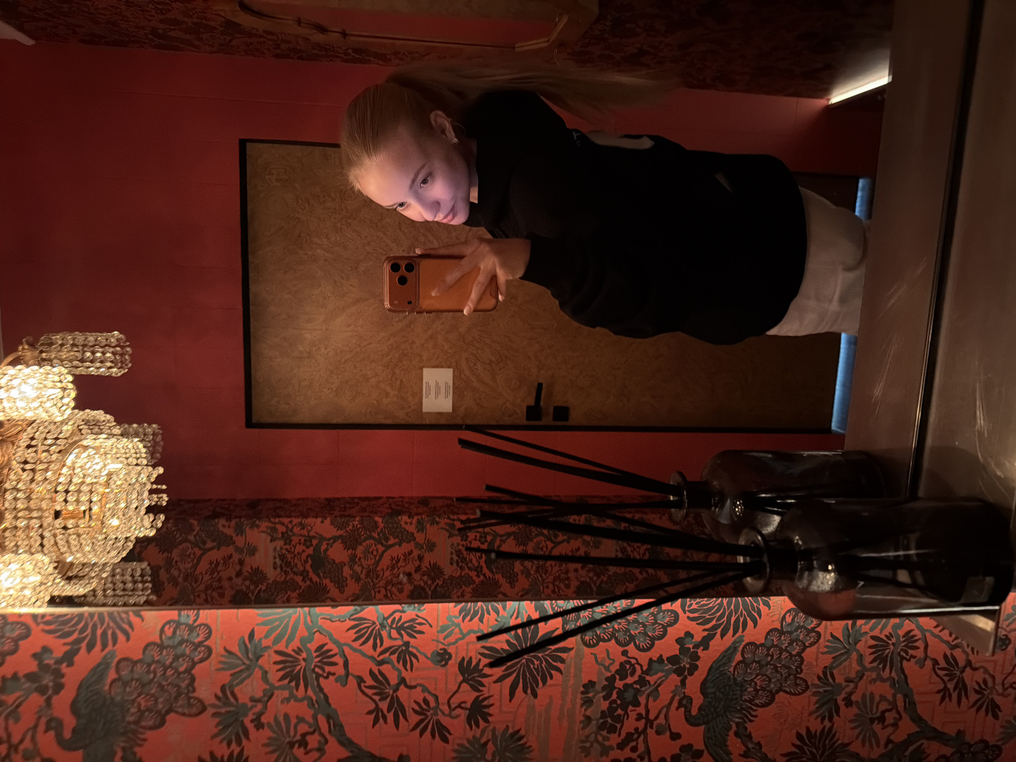 A fair-skinned person with blonde hair in a high ponytail is holding a smartphone horizontally and taking a photo. The person is wearing a black hoodie and light-colored pants. The person is reflected in a mirror. Behind the person's reflection is a door with a rectangular brown panel. The background of the room is red with ornate floral wallpaper near the bottom of the image and a chandelier is present in the lower left. A row of dark brown glass diffusers with black reeds is in the foreground. The location is an unidentified room in Unknown, Unknown.
