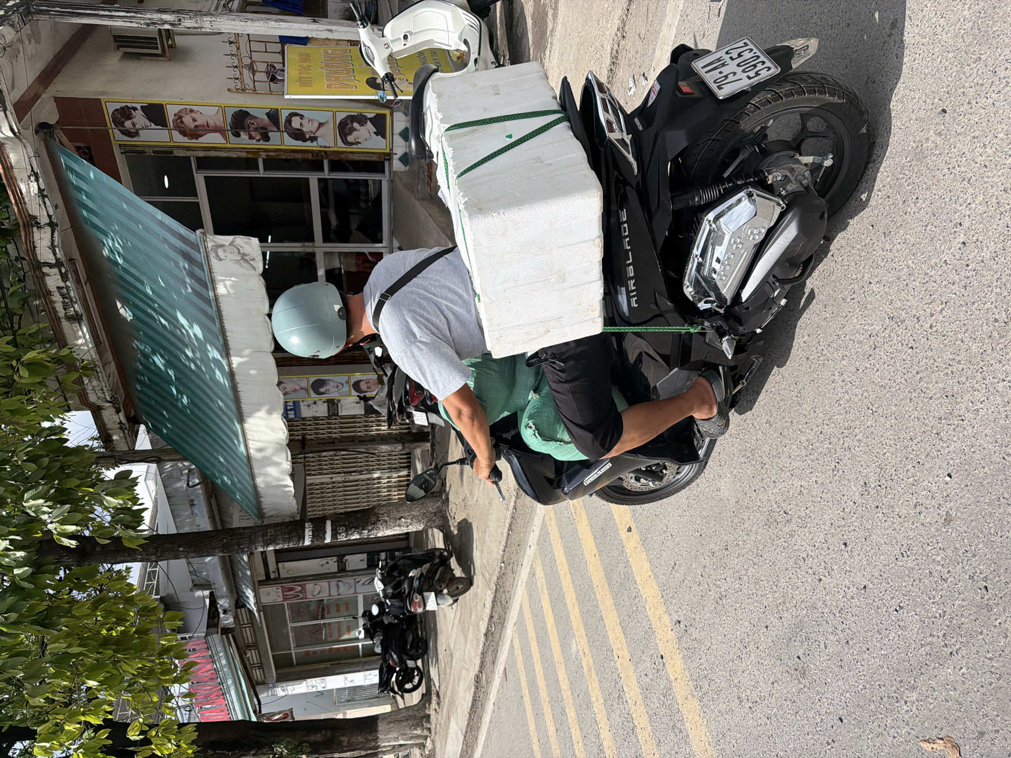 A street scene in Cam Ranh, Vietnam, is captured on a bright, sunny day with strong sunlight casting shadows. The main subject is a person, seen from behind, wearing a light blue helmet, grey t-shirt, and dark shorts or pants, operating a black Honda Airblade scooter. A large white styrofoam box is prominently secured to the back of the scooter with green straps.
The setting appears to be a commercial street, with buildings lining the right side of the road. One building features a green-striped awning and a yellow sign with red lettering. Visible Vietnamese text on this sign includes "PHƯƠNG" and "HỘI TÓC XINH," which translates to "Beautiful Hair Salon." Another partially visible sign shows "BÉ TA." Several headshot images of different hairstyles are displayed below the main sign, indicating a barbershop or hair salon.
Further down the street, another white scooter is parked. A tree with lush green foliage is visible on the left side of the frame, partially obscuring other buildings. The road surface is asphalt, with distinct yellow double lines painted on it. The scooter's license plate, "79-AA 590.52," is clearly visible. The overall impression is one of typical daily activity in a Vietnamese urban environment.