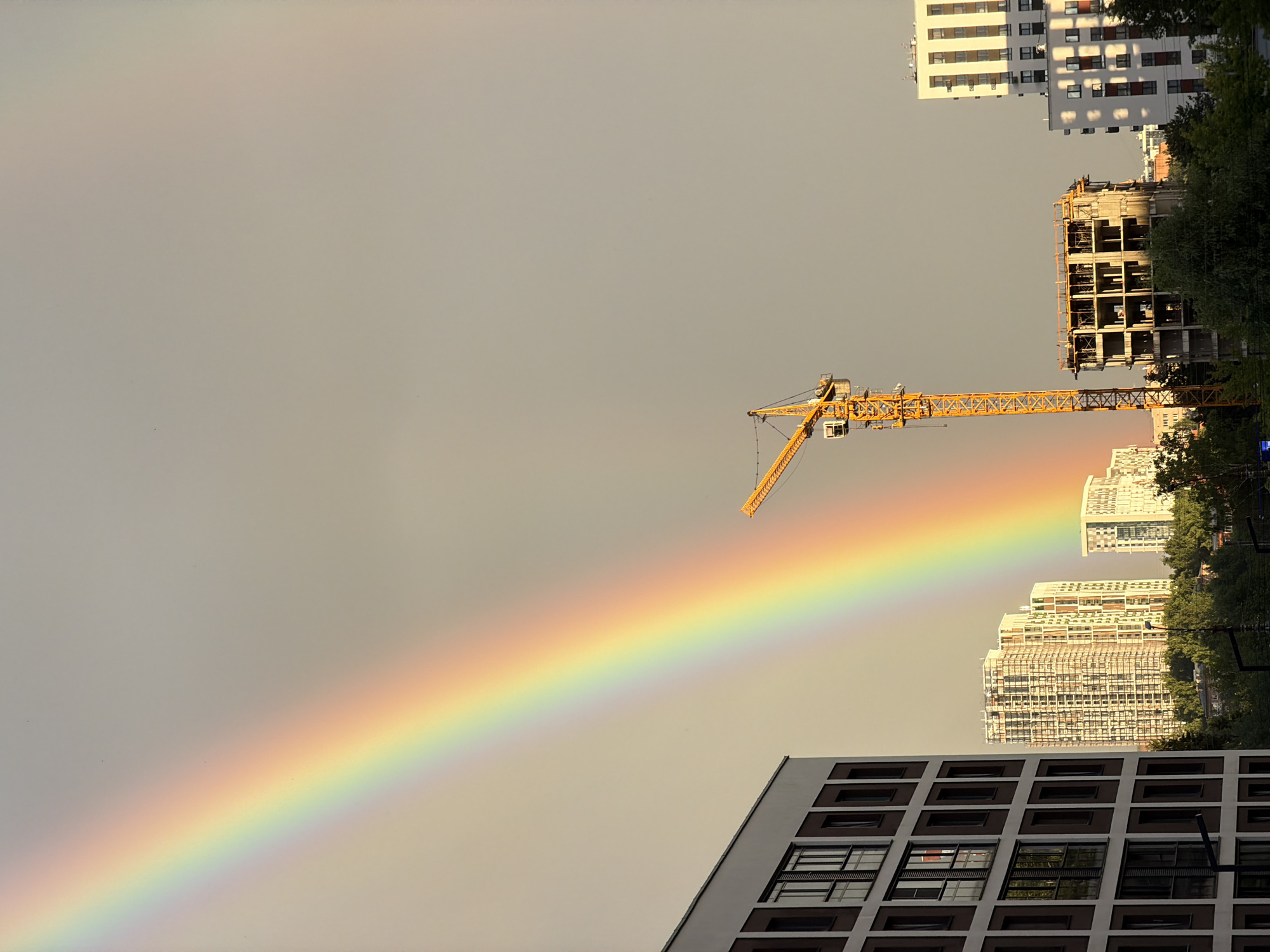The image shows a rainbow stretching across a cloudy sky over a cityscape. In the foreground, to the left, is the side of a modern building with dark brown accents and numerous windows. To the right of the rainbow, a tall yellow construction crane stands prominently. In the middle ground, several buildings are visible, some under construction with exposed rebar and concrete frames, and others appearing to be residential high-rises. The lower part of the image features trees and foliage, suggesting an urban environment with green spaces. The sky is overcast with a subtle gradient, and the light suggests it is either early morning or late afternoon. The scene is likely a residential or mixed-use development area.