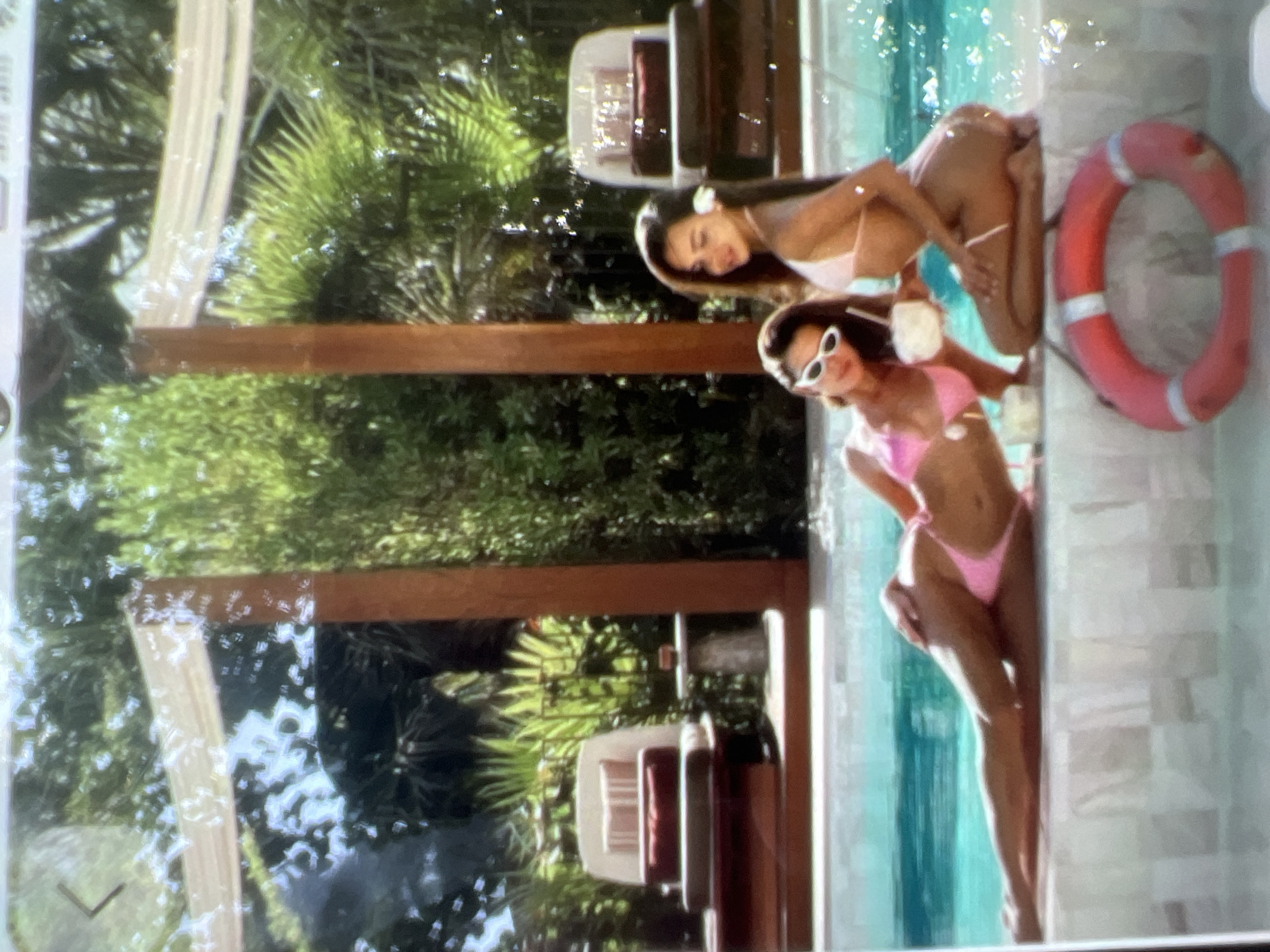 The image shows two women in bikinis by a swimming pool. The woman on the left is lying on the edge of the pool, wearing white sunglasses and a pink bikini. She is holding a white drink. The woman on the right is kneeling and looking at the camera, wearing a pink bikini and a white flower in her hair. She is holding a coconut drink. In the foreground, there is a red life preserver. The background shows lush green foliage and lounge chairs. The scene appears to be a resort or vacation setting. There is no discernible information in the image that suggests the location is in Kara-Dıykan, Kyrgyzstan.