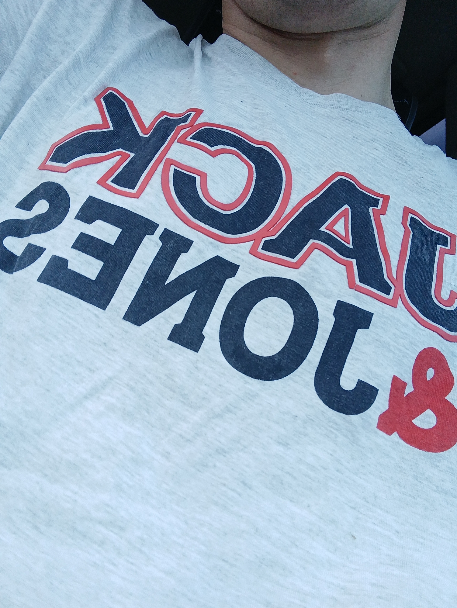 A close-up shot shows the upper chest and neck area of a person wearing a light gray t-shirt with a large graphic print. The graphic features bold, dark blue lettering outlined in red, spelling out "JACK & JONES". The text is angled diagonally across the t-shirt. The person's neck and chin are visible at the top of the frame, and the background is dark and out of focus, suggesting the photo was taken indoors. The overall impression is a casual snapshot of someone wearing branded apparel.